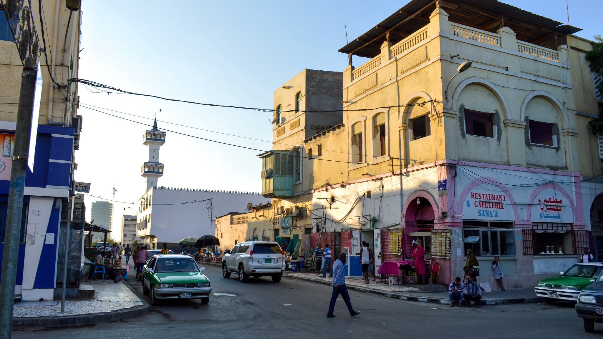 تظهر الصورة مشهداً لحياة الشارع في مدينة، حيث تتواجد المباني ذات الطراز المعماري التقليدي ذو الألوان الفاتحة. في الخلفية، يمكن رؤية مئذنة مسجد تُشير إلى وجود مكان للصلاة. في الشارع، تُظهر الحركة بعض المارة والسيارات، مما يعطي انطباعاً بالحيوية والنشاط. المناطق المحيطة بالأبنية تبدو مزدحمة، مع وجود عدد من المحلات التجارية والمقاهي. السماء زرقاء، مما يوحي بأن الوقت هو فترة الصباح أو بداية النهار.