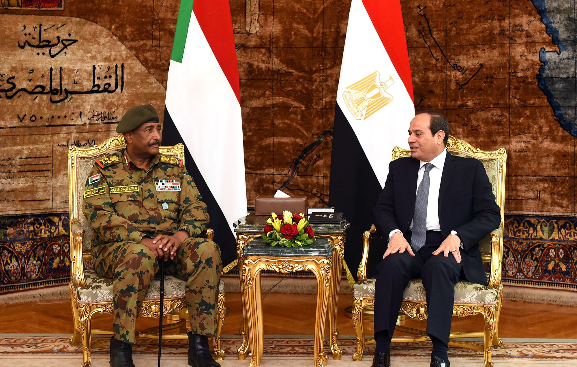 تظهر الصورة لقاءً رسميًا بين شخصين يجلسان على كراسي فاخرة أمام علمي السودان ومصر. خلفهم يوجد ستار مزخرف يضيف لمسة فخمة للمكان. الشخص على اليسار يرتدي زيًا عسكريًا ويبدو أنه مسؤول عسكري، بينما الشخص على اليمين يرتدي بدلة رسمية. يعبر كلا الشخصين عن تواصل دبلوماسي بين البلدين.