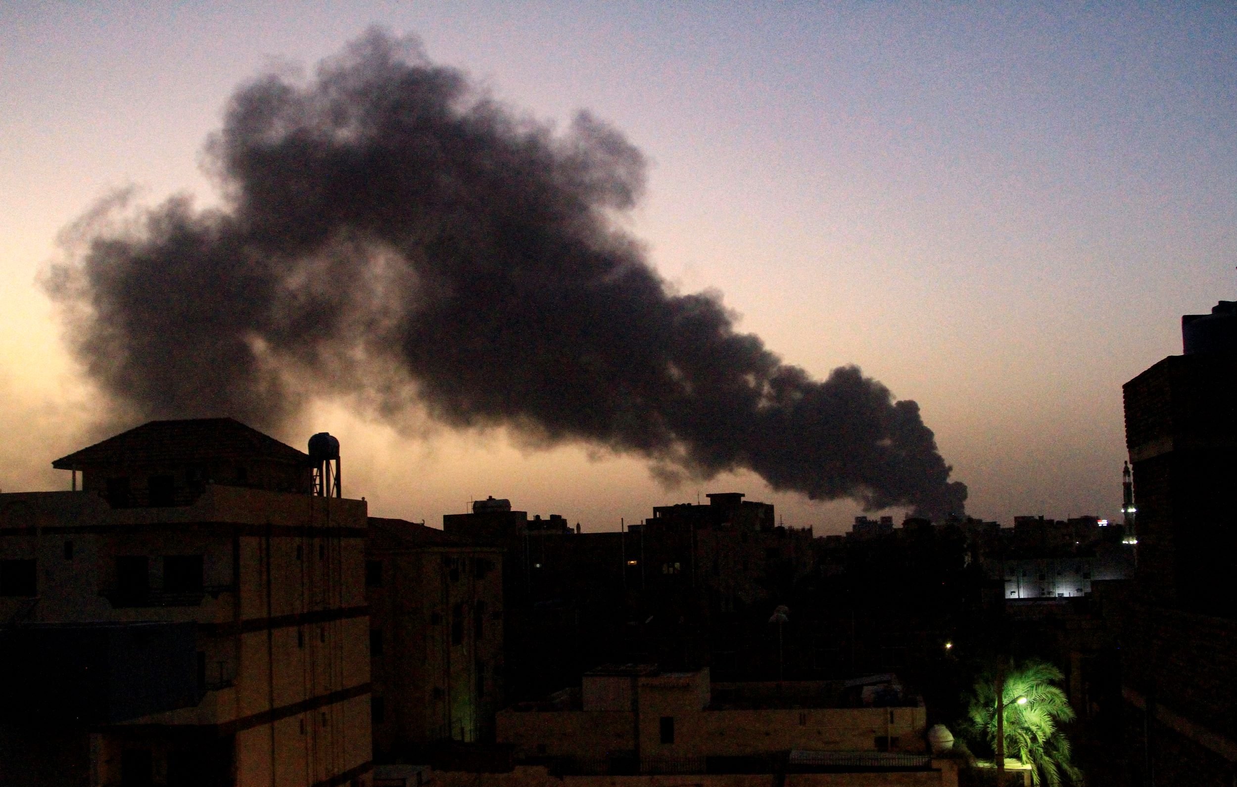 تظهر الصورة مشهداً لسماء غامقة تتخللها غيوم من الدخان الأسود تتصاعد في الأفق. تبدو المباني في المقدمة بلون داكن، مما يعكس ظلام الوقت الذي قد يكون عند الغسق. بينما تضفي أشجار النخيل لمسة خضراء في الجانب. الدخان يبدو كثيفاً وواضحاً، مما يشير إلى وجود حريق أو حدث صناعي في المنطقة.