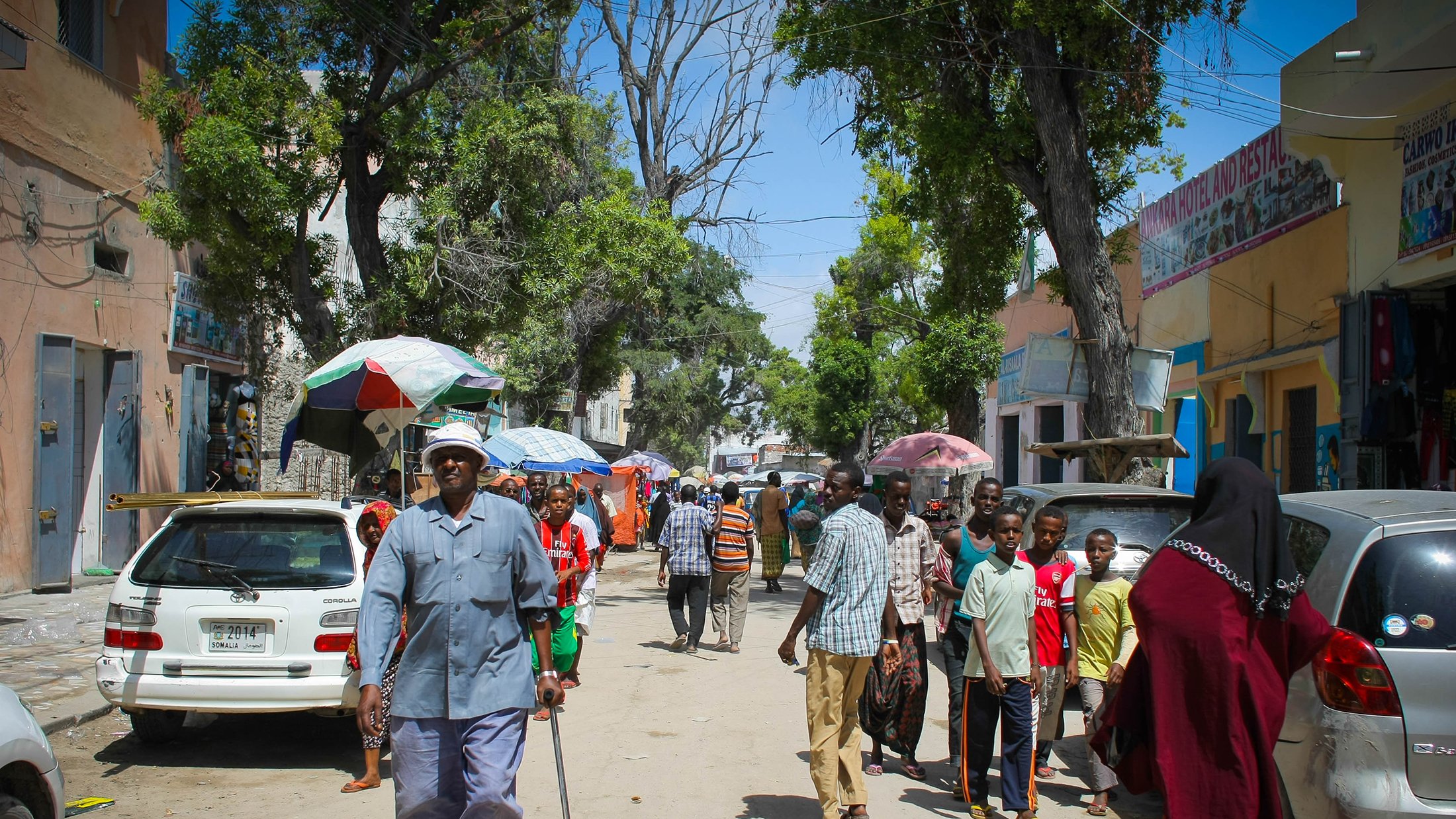 تظهر الصورة شارعا في منطقة حيوية، حيث يسير مجموعة من الأشخاص في اتجاهات مختلفة. في المقدمة، يوجد رجل مسن يستخدم عكازا للمشي، بينما يتواجد أشخاص آخرون في الخلفية، بعضهم يتحدث والبعض الآخر يسير. هناك سيارات متوقفة على الجانبين، وتظلل الأشجار والشمس المكان. يبدو أن الجو مشمس والناس يقومون بأعمالهم اليومية.