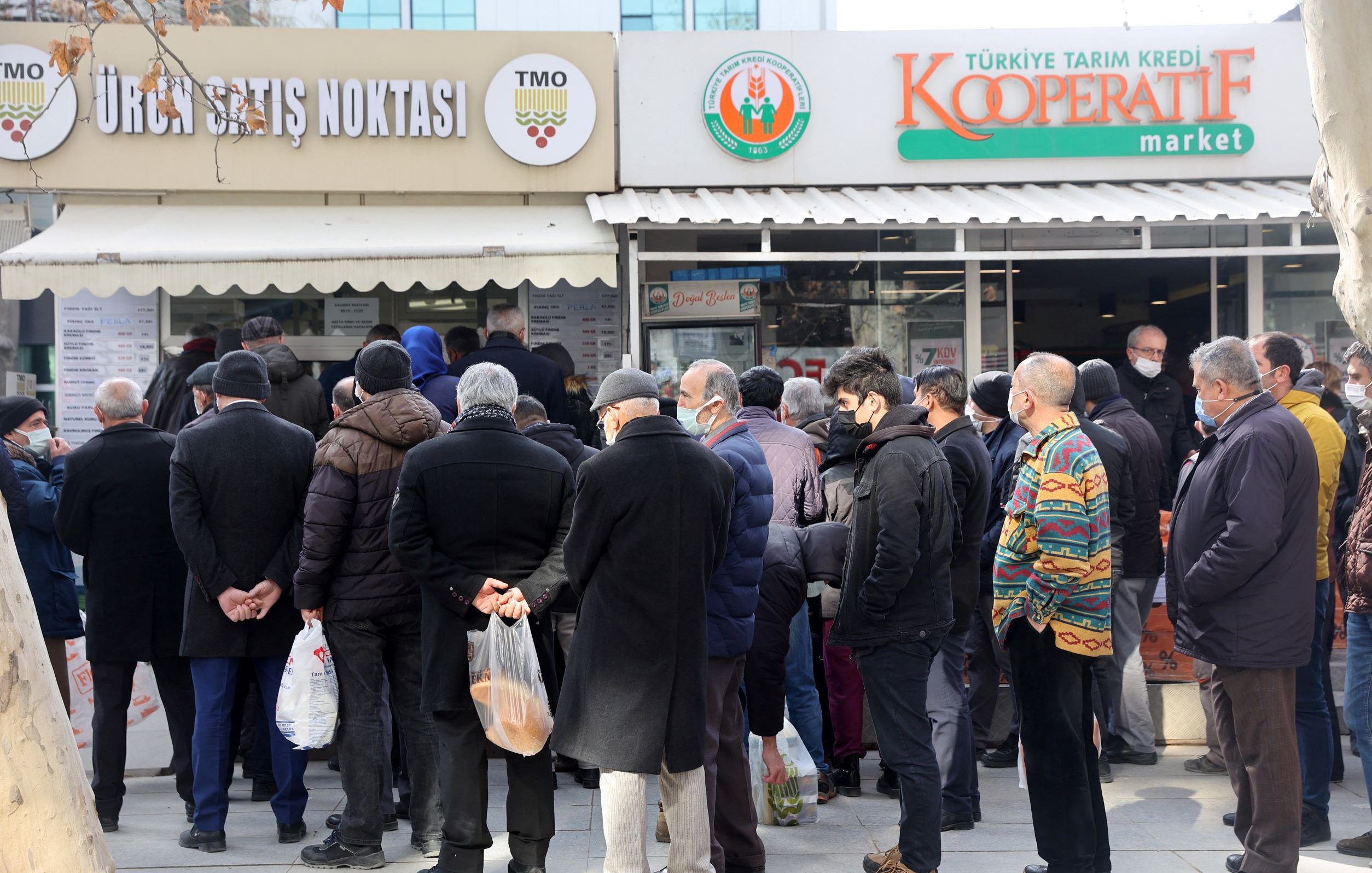 الصورة تظهر مجموعة من الأشخاص الواقفين في طابور أمام متجر يُظهر لافتة تدل على أنه نقطة بيع للمنتجات. يبدو أن معظم الأشخاص يرتدون ملابس شتوية وأنهم يحملون أكياسًا. الجو يبدو هادئًا، مع وجود أشجار حول المكان، بينما يبدو أن بعضها قد فقد أوراقه بسبب البرد. المتجر يحمل اسم "كوoperative" ويبدو أنه يبيع مواد غذائية أو منتجات محلية.