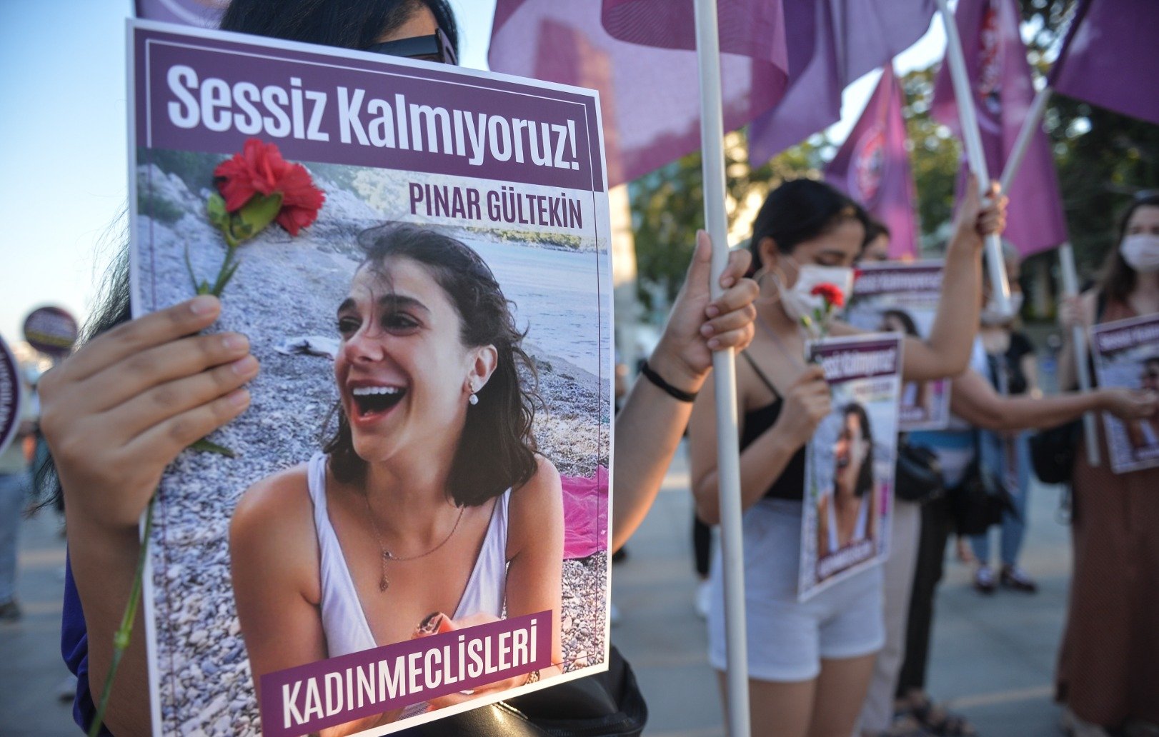 تظهر الصورة مجموعة من النساء أثناء تظاهرهن. تحمل إحداهن لافتة مكتوب عليها "Sessiz Kalmıyoruz!" (لا نصمت!) وصورة لامرأة تُدعى "Pınar Gültekin". كما تحمل إحدى المتظاهرات زهرة حمراء، مما يشير إلى أن المظاهرة تتعلق بقضية اجتماعية أو حقوق النساء. يُظهر المشهد روح التضامن والمطالبة بالعدالة.