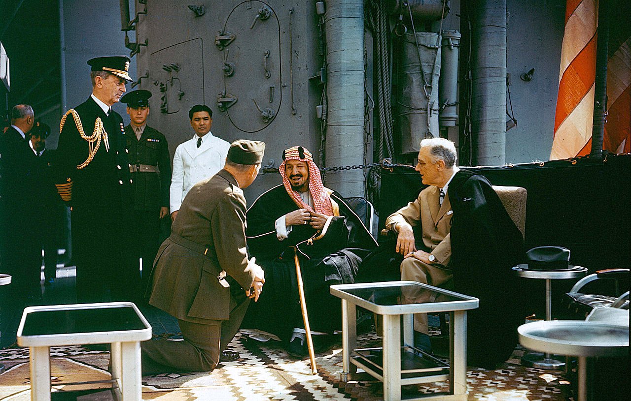 تظهر الصورة مجموعة من الرجال في إطار رسمي. يجلس رجل يرتدي ملابس تقليدية على كرسي، بينما يوجد أمامه رجل آخر يرتدي زيًا عسكريًا، يبدو أنه يتحدث إليه أو يقدم له تحية. خلفهم، يظهر عدد من الرجال يرتدون بدلات رسمية، وبعضهم ينظر نحو الجالسين. يبدو المشهد وكأنه حدث تاريخي أو اجتماع مهم، حيث يعبر تعبير وجه الرجل الجالس عن ارتياح أو سرور.