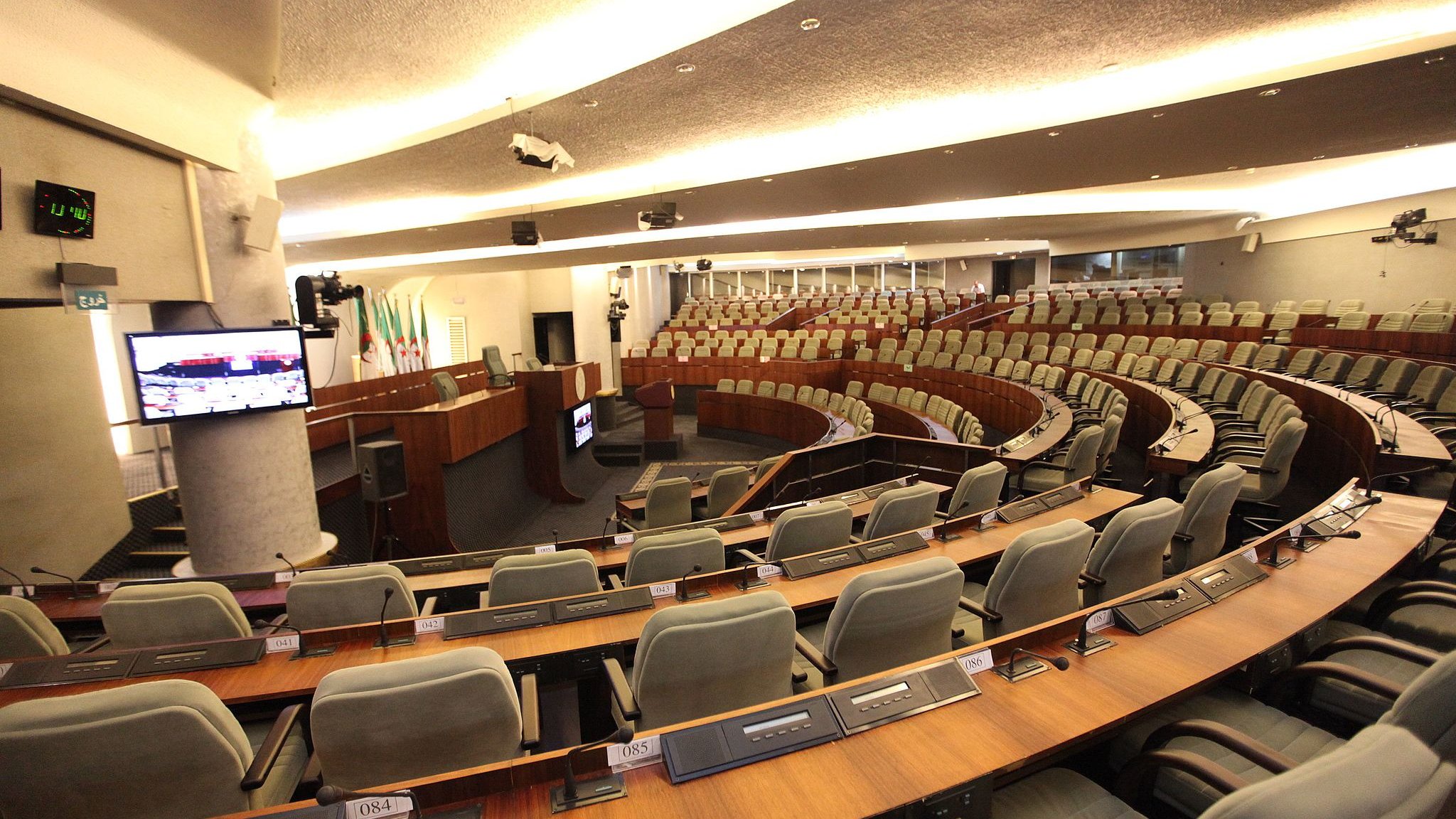 تظهر الصورة قاعة اجتماعات واسعة، تتميز بتصميم حديث ومرتب. تحتوي القاعة على مقاعد مريحة مرتبة في صفوف منحدرة، مما يتيح للمتحدثين رؤية الجمهور. يوجد في الخلف منصة للمتحدثين، مع معدات سمعية بصرية مثل الشاشات والأجهزة الصوتية. الألوان المستخدمة في الديكور تمزج بين الألوان الخشبية الفاتحة والبيضاء، مما يعطي مظهراً أنيقاً وعصرياً.