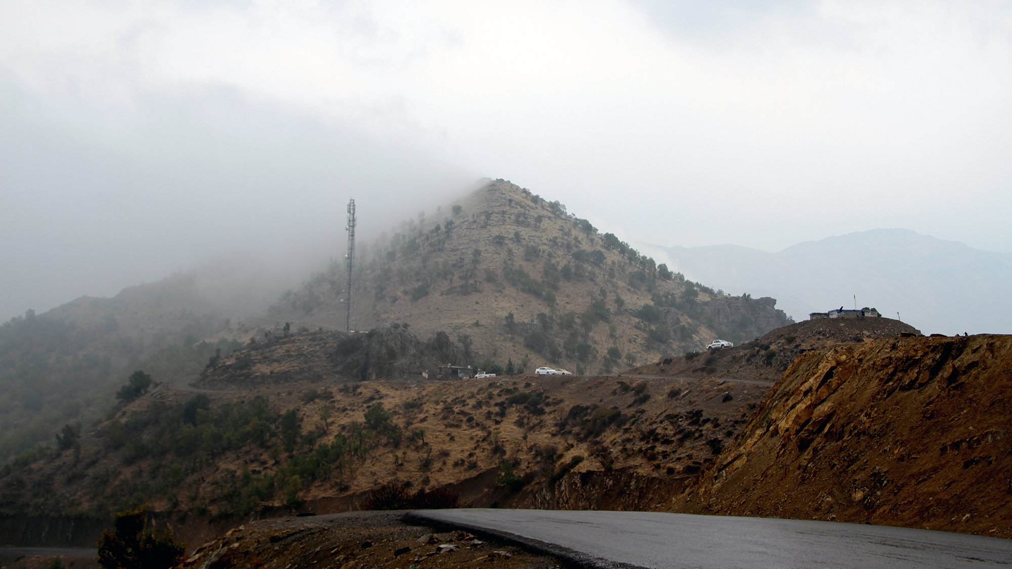 تظهر في الصورة مشهدًا جبليًا يكتنفه الضباب، حيث تتساقط السحب بشكل كثيف على الجبل. في الجزء العلوي من الجبل، يمكن رؤية برج اتصالات. الطريق يلتف حول الجبل، ويظهر بشكل مائل مع اختلافات في الألوان بين الصخور والتربة. الأجواء تبدو هادئة، مع لمسات من الغموض بسبب الضباب.