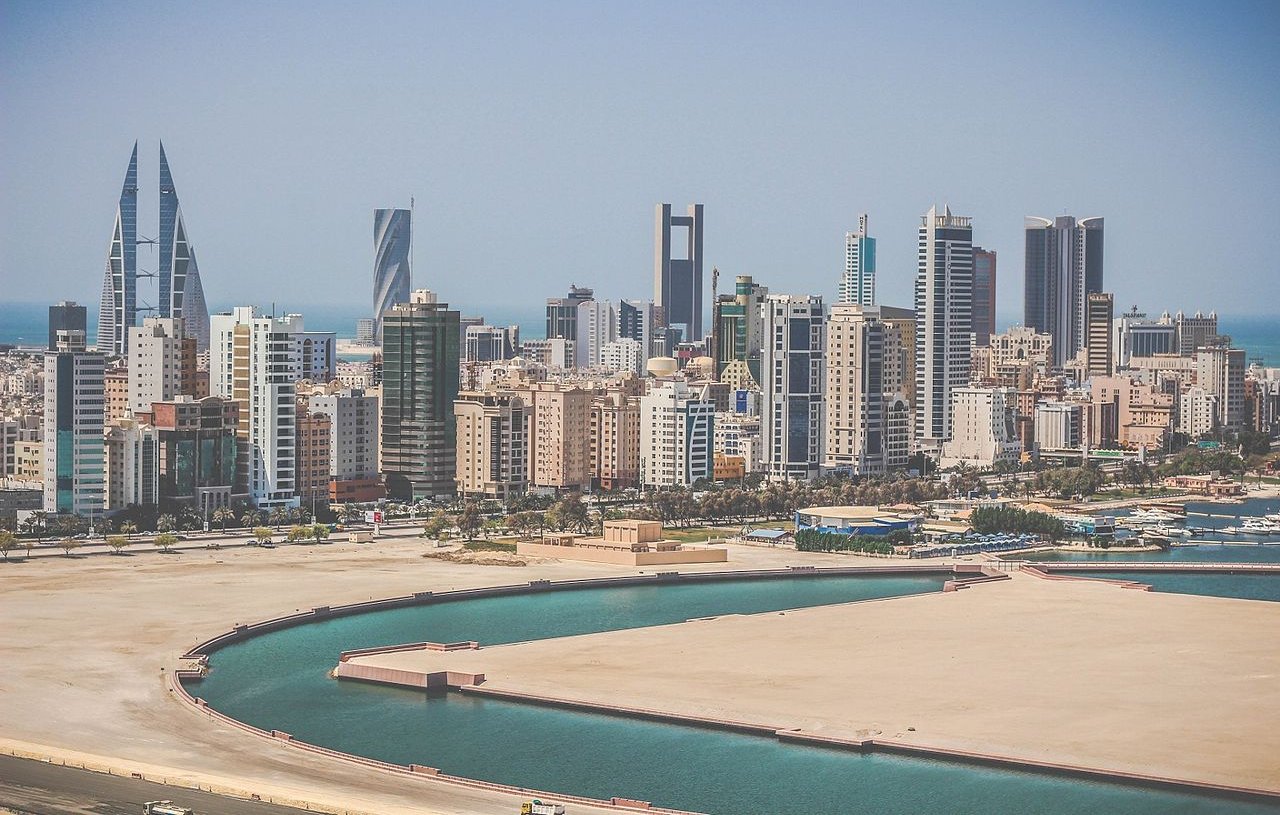 تظهر الصورة مشهداً لمدينة عصرية تضم العديد من ناطحات السحاب والمباني الحديثة. يتواجد في الخلفية مجموعة من الأبراج المميزة، بعضها ذو تصميم فريد. كما يظهر جزء من البحر على اليمين، مع مرفأ صغير يضم قوارب. السماء تبدو صافية وتضيء بأشعة الشمس، مما يضفي شعوراً بالوضوح والنشاط على المشهد العام.