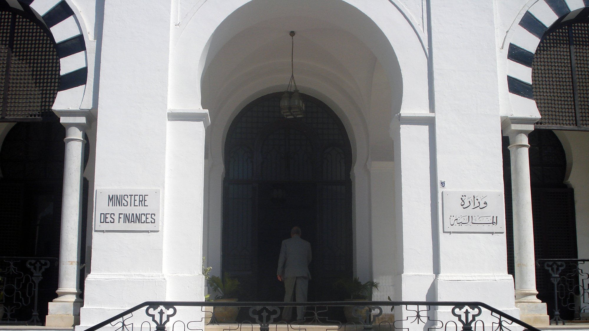 تظهر الصورة مدخل مبنى حكومي يُعرف باسم "وزارة المالية". يتميز المبنى بتصميم معماري جذاب، حيث يمكن رؤية الأبواب الكبيرة والنوافذ المزخرفة. المدخل محاط بأعمدة وأقواس، مما يعطيه طابعاً تقليدياً. هناك لافتات على الجانبين، واحدة باللغة الفرنسية والأخرى بالعربية، تشير إلى اسم الوزارة. يُلاحظ أيضاً شخص يقف أمام المدخل، مما يعكس النشاط في هذا المكان.