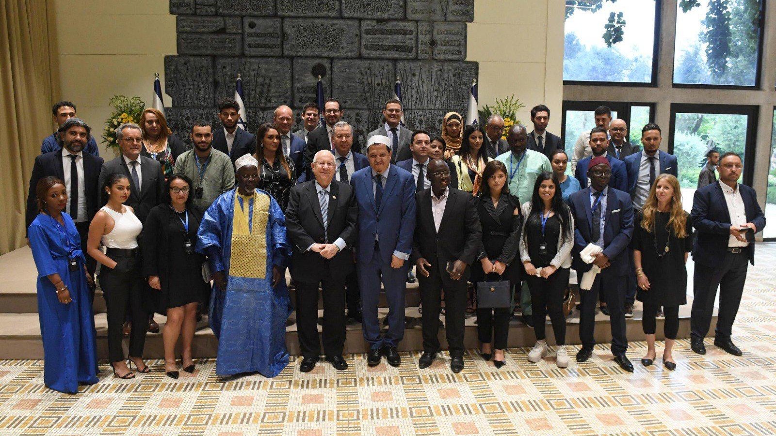 تظهر الصورة مجموعة كبيرة من الأشخاص الذين يقفون معاً في مكان مغلق. يرتدي البعض ملابس رسمية، بينما يرتدي الآخرون ملابس تقليدية. يوجد خلفهم علم أو شعار يشير إلى وهو مرتكز على جدار مزخرف. يبدو أن الحدث يجمع أفراداً من خلفيات متنوعة، ويمثل مجموعة من المشاركين في مناسبة رسمية أو مؤتمر. تعكس الصورة تفاعلًا إيجابيًا بينهم.