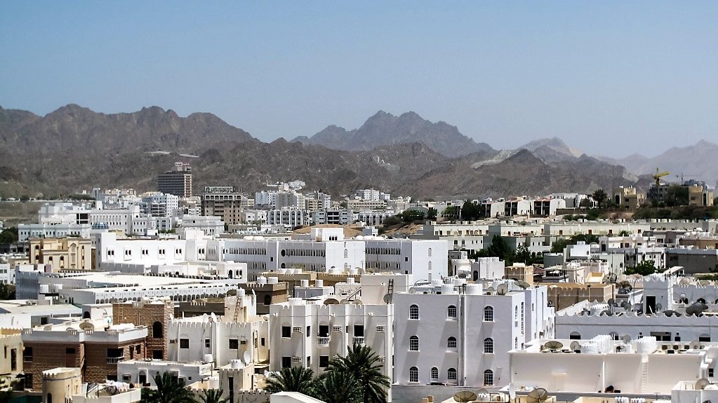 الصورة تُظهر مشهداً لمدينة محاطة بالجبال، حيث تتداخل المباني البيضاء مع بعض اللمسات الحديثة. تمتاز rooftops بأسقفها المسطحة، بينما تظهر في الخلفية الجبال الشاهقة التي تضفي جمالاً على المنظر. السماء نقية وزرقاء، مما يضيف إحساساً بالصفاء والهدوء للمنطقة.