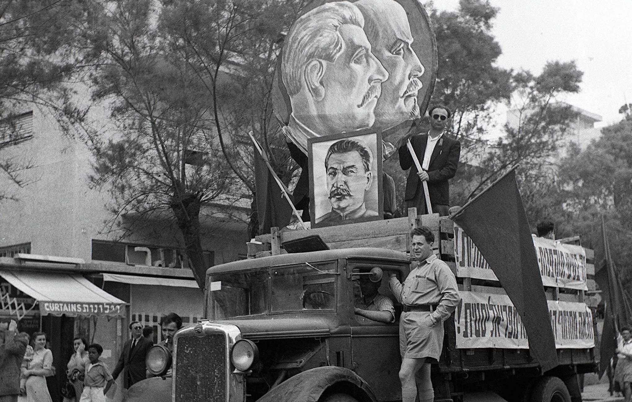 تظهر الصورة عربة نقل تحمل لافتة ضخمة تضم صورًا لشخصيات تاريخية، يبدو أن لها دلالات سياسية. يتواجد شخصان على العربة، أحدهما يرتدي زيًا مميزًا. تتوسط العربة مجموعة من الأعلام، ويبدو أن هناك تجمعًا من الناس في الخلفية، مما يشير إلى حدث جماهيري أو مسيرة. المشهد يعبّر عن أجواء زمنية معينة قد تكون مرتبطة بفترة تاريخية أو مناسبة سياسية.