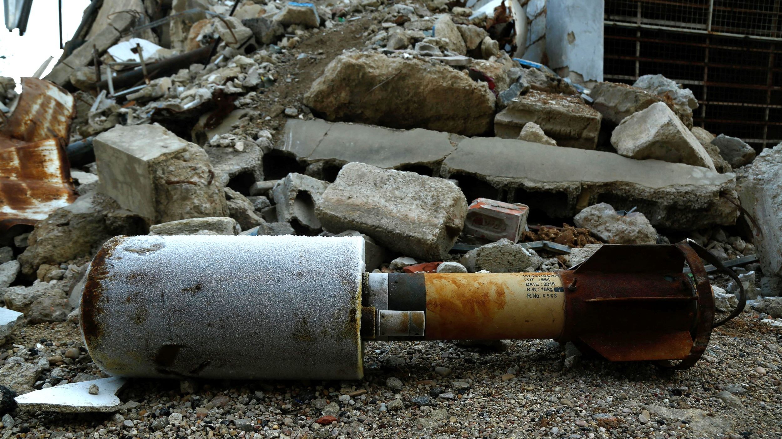 الصورة تظهر قنبلة أو جسم متفجر ملقى على الأرض، محاط بأحجار وأنقاض تشير إلى تدمير في المنطقة. يبدو أن الساحة غير مرتبة، والأرض مليئة بالردم والحطام، مما يشتت الانتباه عن الجسم المتفجر. يظهر أن هذا المكان شهد حدثًا مروعًا أدى إلى تدمير كبير.