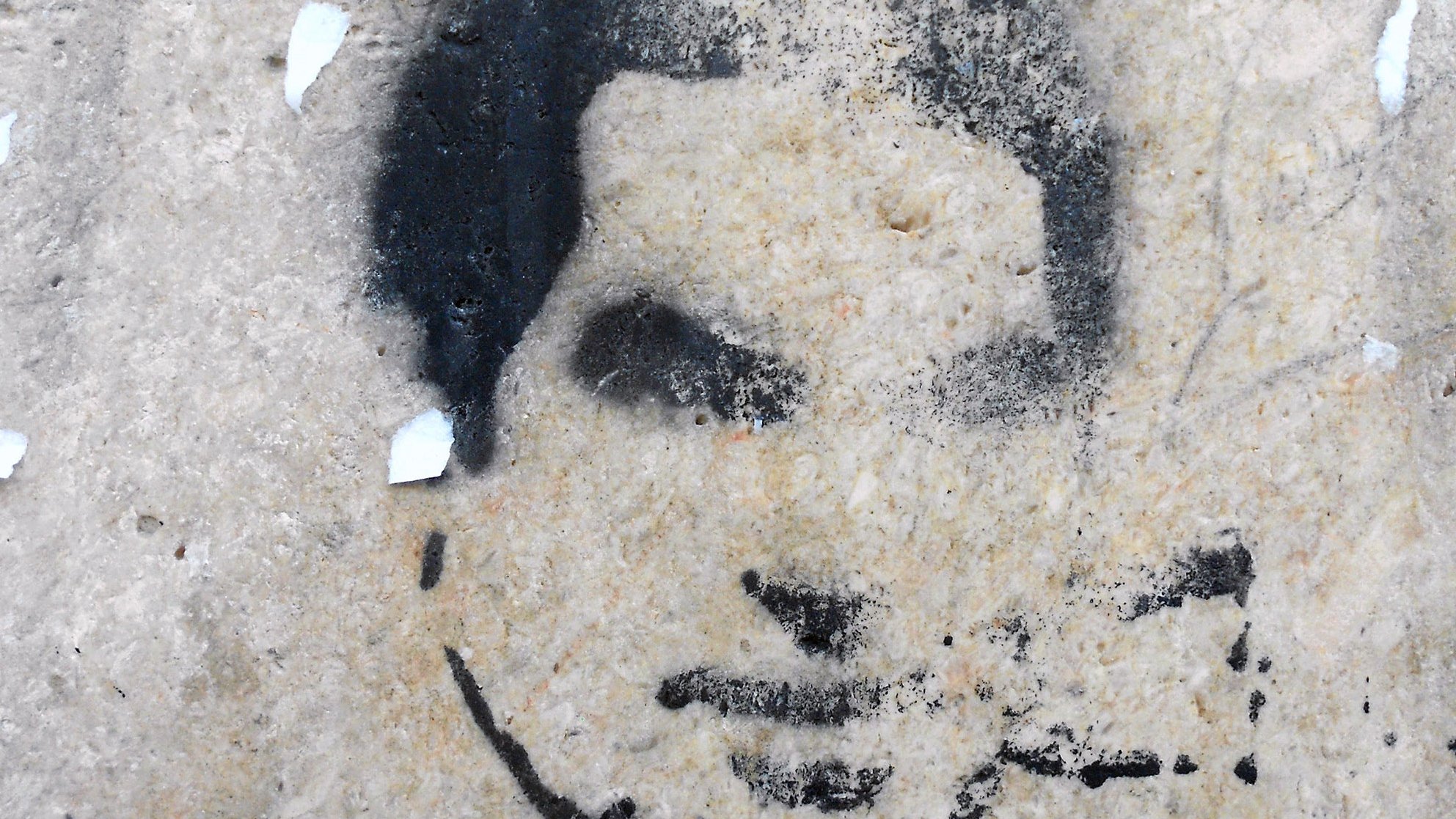 الصورة تظهر رسومًا جدارية تُظهر وجه إنسان بأسلوب الطلاء الرش. يبدو أن الرسم مقطوع بعض الشيء، مع تفاصيل غامضة لفم وأنف وعينين. الخلفية غير منتظمة وقد تشتمل على بعض آثار الندوب أو التآكل، مما يعطي الانطباع بأنها قد رسمت على سطح قديم أو متآكل. اللون المستخدم في الرسمة أسود بينما الخلفية ذات لون فاتح.