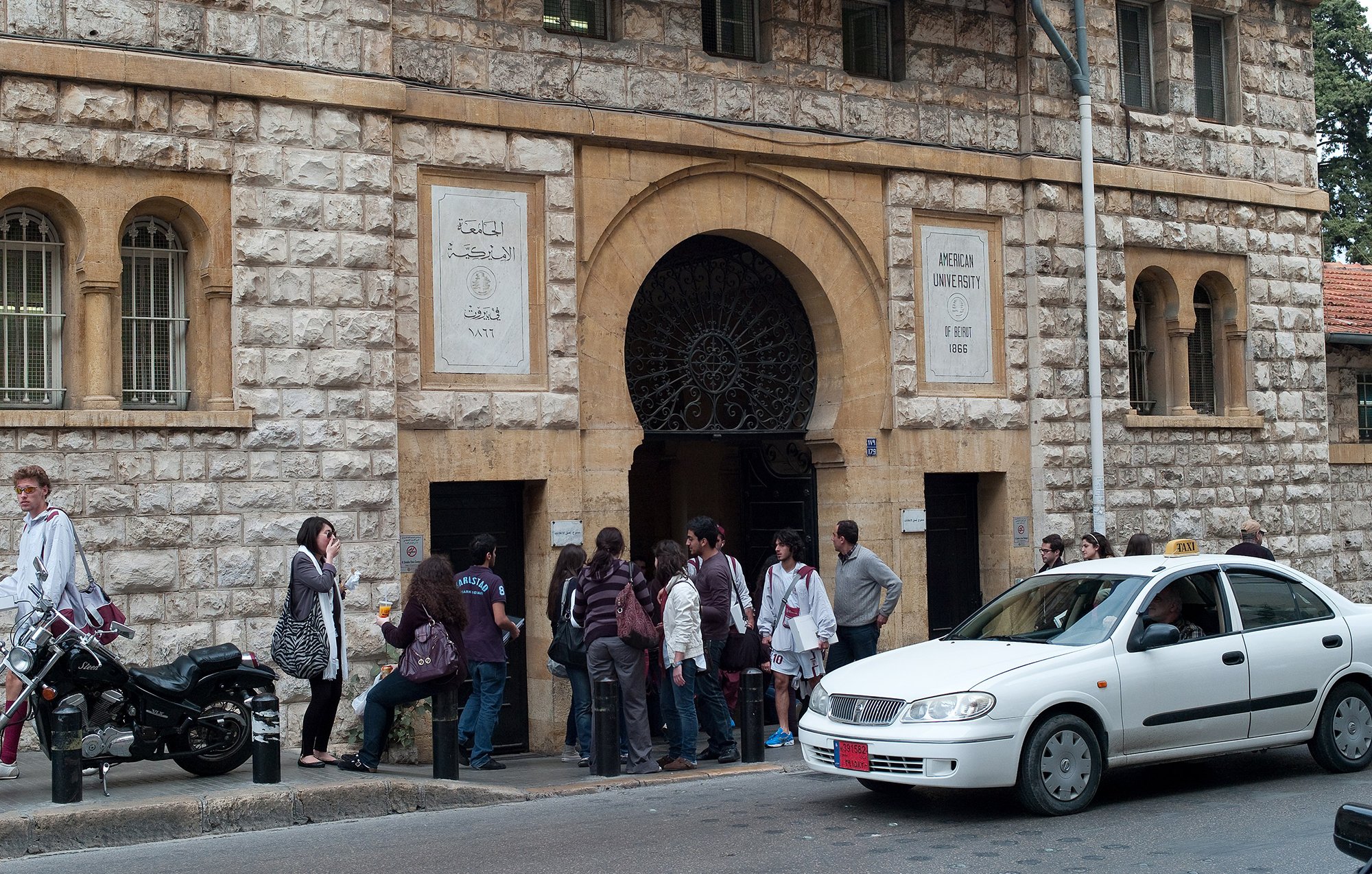 الصورة تظهر مدخل جامعة معمارية مميزة، حيث يمكن رؤية واجهة المبنى التي تتكون من حجر باهت وأعمدة مميزة. أمام المدخل، يوجد مجموعة من الطلاب أو الأشخاص في صف، مما يدل على أنهم قد يكونون في انتظار التسجيل أو الدخول. إلى جانب المدخل، توجد دراجة نارية ومركبة تسير على الطريق، مما يعطي انطباعاً عن الحياة اليومية والحركة في المنطقة.背景وضح صورة جوًا من النشاط والاهتمام بالتعليم.