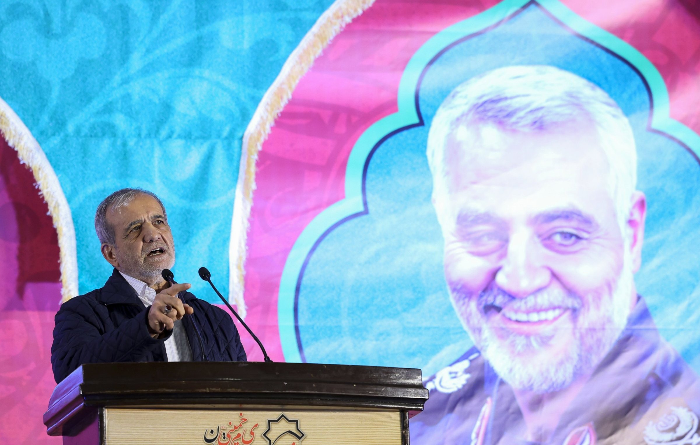 The image features a man speaking at a podium, with a colorful backdrop that includes a large portrait of another individual smiling. The background has a decorative design, predominantly featuring blue and pink colors. The speaker appears animated and engaged, and the setting suggests it may be a formal event or speech, possibly commemorating or honoring the person depicted in the portrait.
