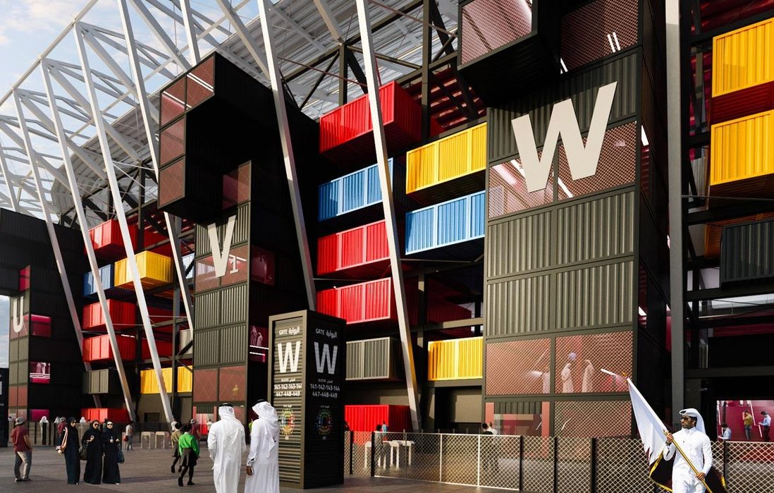 الصورة تظهر تصميمًا عصريًا لمنطقة بجانب ملعب أو منشأة رياضية. يتميز التصميم بألوان زاهية تتضمن الصناديق الملونة بالأسود والأحمر والأصفر والأزرق. هناك أيضًا مدخل واسع مع شعارات واضحة. في الجزء الأمامي، يظهر بعض الأشخاص يرتدون الملابس التقليدية، مما يشير إلى إمكانية وقوع الحدث في منطقة عربية أو إسلامية. السماء تتسم بصبغة رمادية توحي بوقت الغسق.