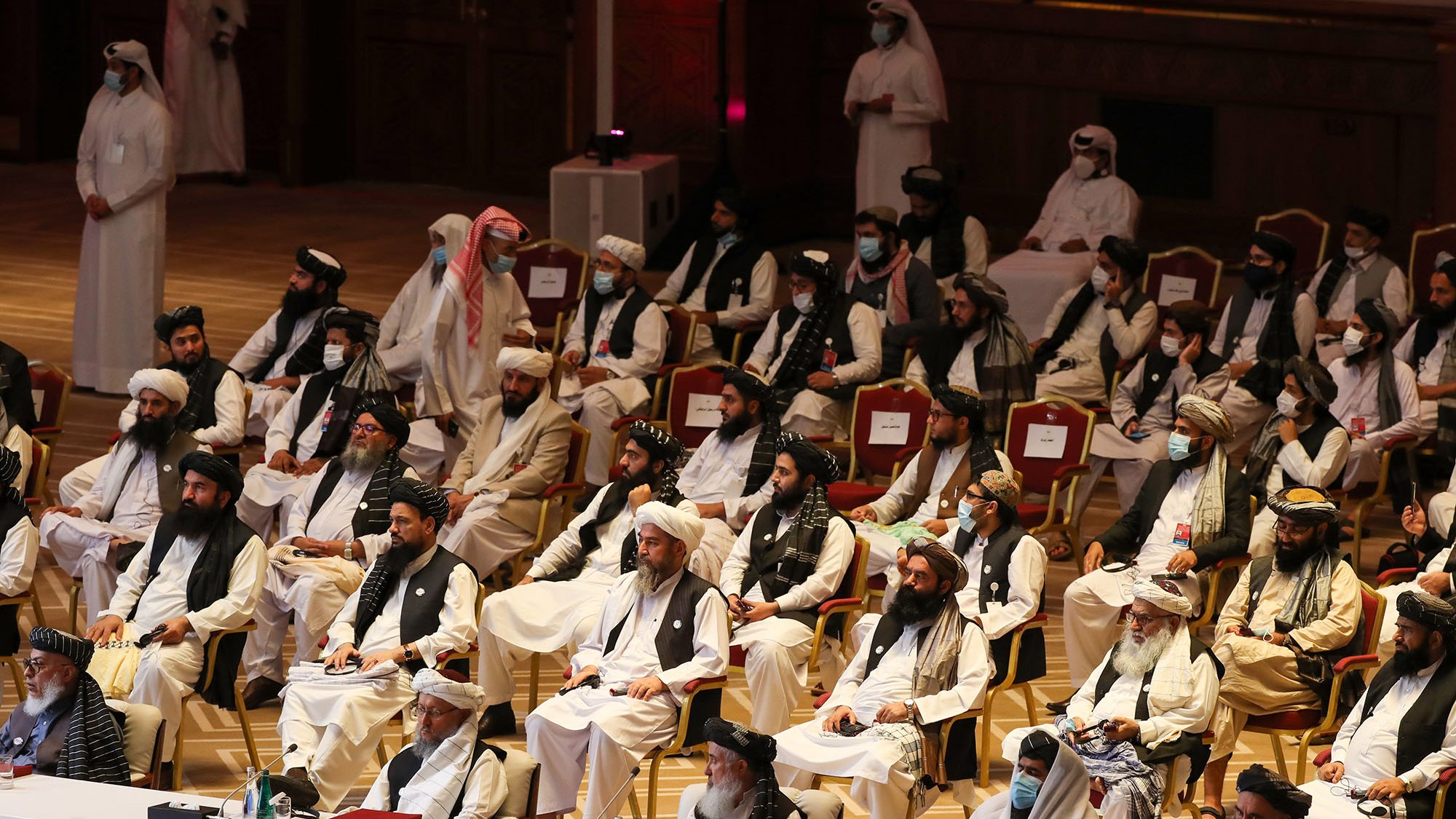 تظهر الصورة مجموعة من الأشخاص يجلسون في قاعة كبيرة، يرتدون ملابس تقليدية. يبدو أن بعضهم يرتدي الأشرطة والعمائم، بينما يحضر آخرون بزي رسمي. القاعة مزينة بروعة، وهناك تفاصيل معمارية خلفهم، مما يشير إلى أنها حدث رسمي أو مؤتمر. يبدو أن بعض الحضور يرتدون كمامات، مما يدل على الاحتياطات الصحية.