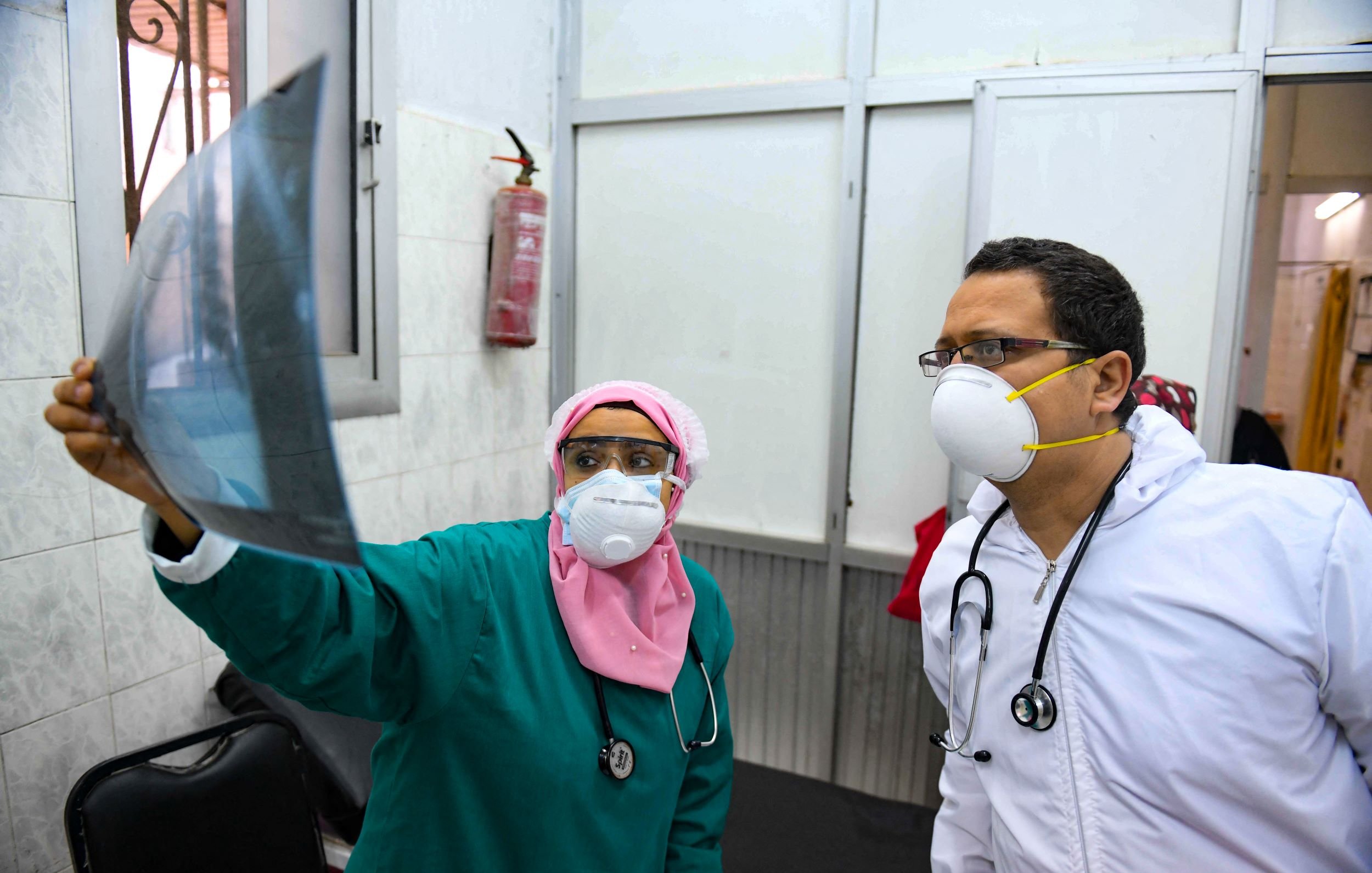 تظهر في الصورة مهنية طبية ترتدي زيًا طبيًا أخضر وغطاء رأس ورديًا، وتفحص صورة أشعة. إلى جانبها، يوجد طبيب يرتدي سترة بيضاء وقناع وجه وقناع تنفس. كلاهما يبدوان مهتمين بالمعلومات الموجودة في صورة الأشعة، ويظهر في الخلفية جدار مكتبي ومعدات طبية.