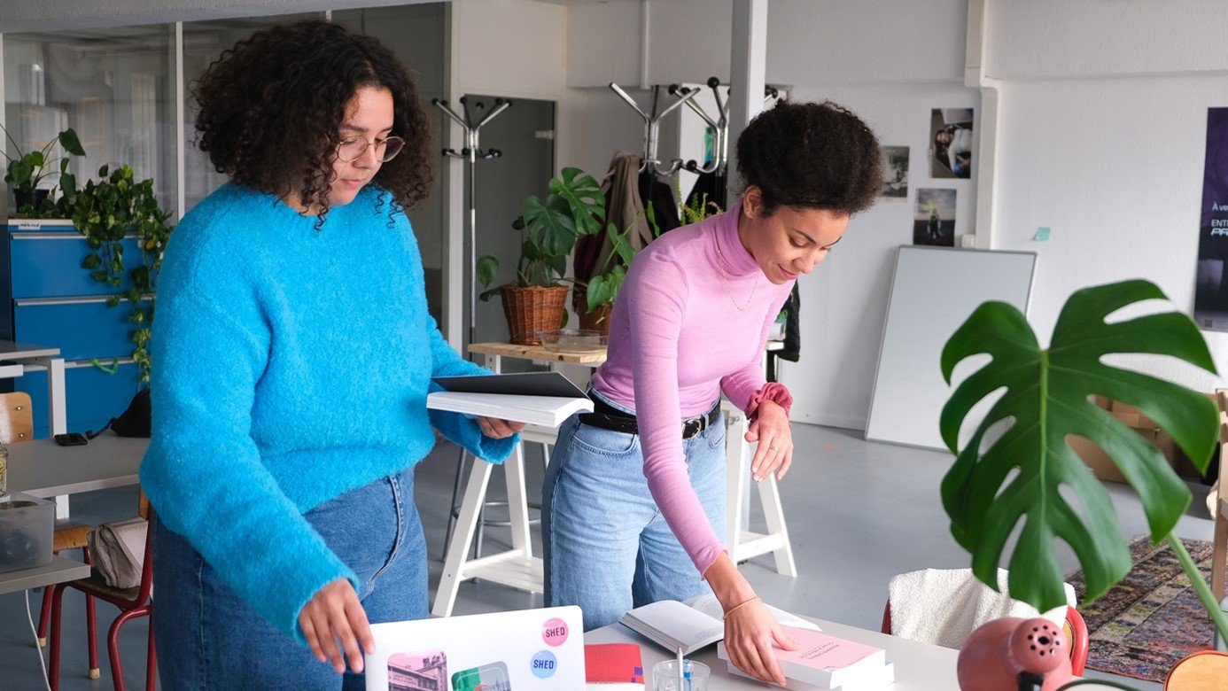 في الصورة، تظهر امرأتان يعملان معًا في مكتب عصري. إحدى السيدتين ترتدي سويتر زرقاء ومرفع، بينما الأخرى ترتدي سويتر وردي وبنطلون جينز. يبدو أنهما مشغولتان بتنظيم بعض الملفات أو الكتب على الطاولة. يوجد على الطاولة كمبيوتر محمول وبعض المقتنيات مثل الكوسات والكتب والمجلة. الخلفية تحتوي على نباتات وأثاث بسيط، مما يعكس جوًا من الإبداع والتعاون.