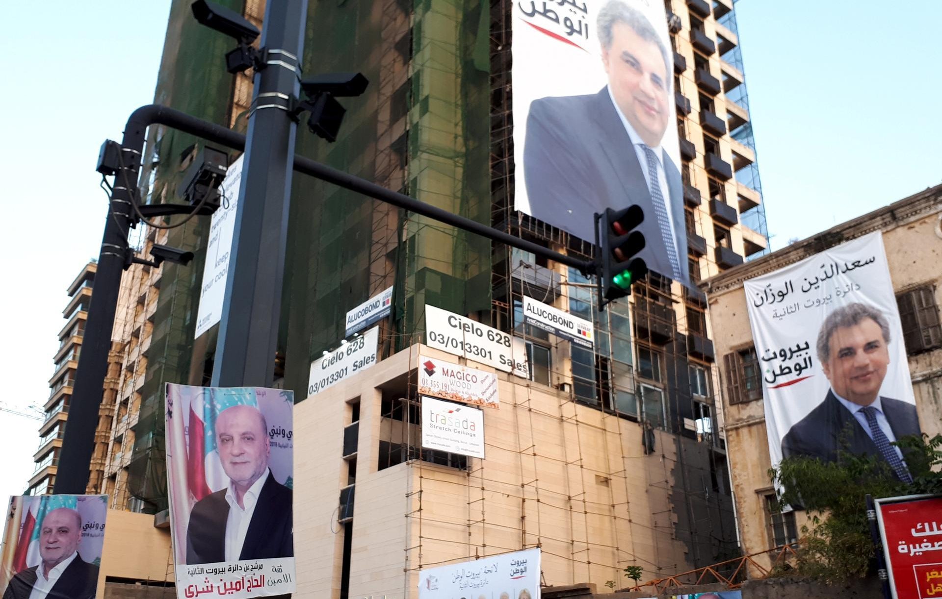 تظهر في الصورة واجهة مبنى يتوسطها لافتات كبيرة تحمل صورًا وحملات انتخابية لأشخاص مختلفين. هناك أيضًا لافتات صغيرة مُعلقة في الأسفل، بالإضافة إلى إشارات المرور، مما يدل على أن المكان هو شارع في منطقة حضرية. المبنى يظهر عليه علامات التجديد أو الصيانة. تعكس الصورة أجواء الانتخابات والنشاط السياسي في المنطقة.