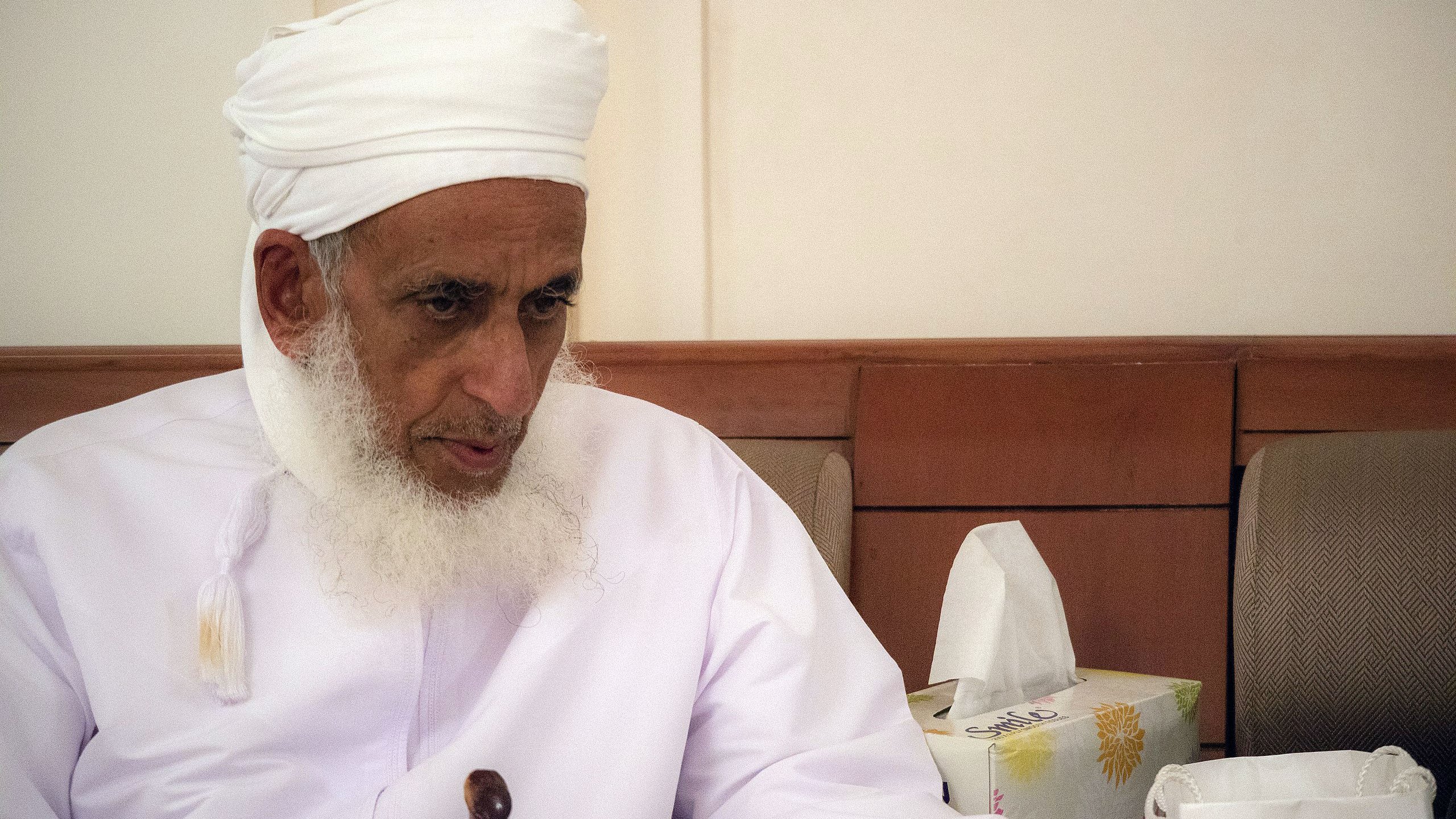 تظهر في الصورة رجل مسن يجلس في مكان مغلق. يرتدي روبًا تقليديًا أبيض مع عمامة بيضاء، ويبدو عليه الوقار والحكمة. لديه لحية بيضاء طويلة، وينظر إلى شيء ما أمامه بتركيز. بجانبه توجد علبة مناديل ورقية، مما يوحي بأنها بيئة اجتماعية أو مريحة. تعكس تعابيره احترامه وتفكيره العميق.