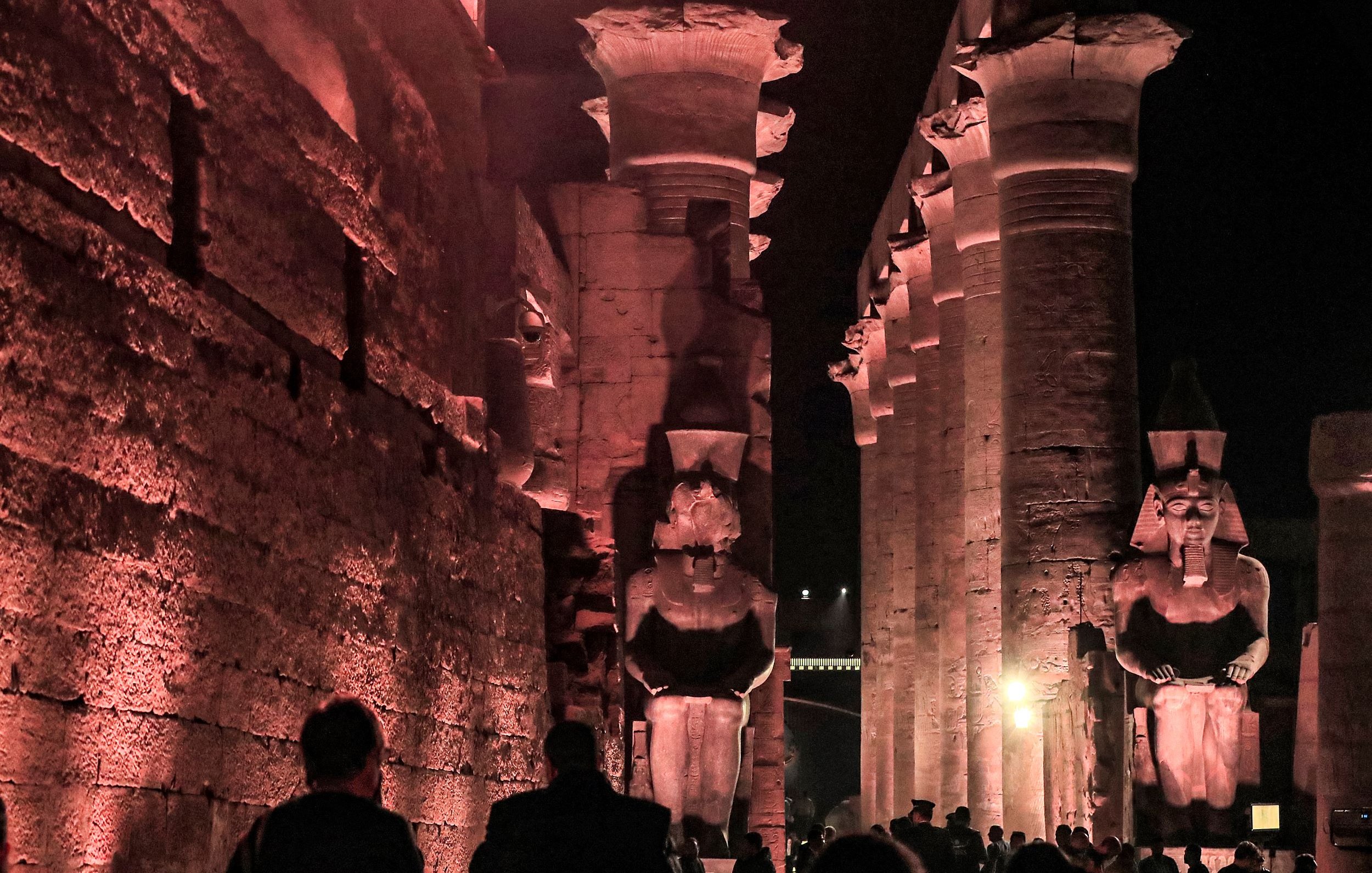 الصورة تظهر معالم أثرية قديمة، حيث يمكن رؤية أعمدة ضخمة ومعمار تاريخي فاخر. الإضاءة الليلية تضفي جوًا سحريًا على المكان، مع تماثيل كبيرة تشكل جزءًا من التصميم المعماري. يبدو أن هناك زوارًا يستكشفون هذا الموقع التاريخي، مما يدل على شهرة وأهمية هذا المكان السياحي.