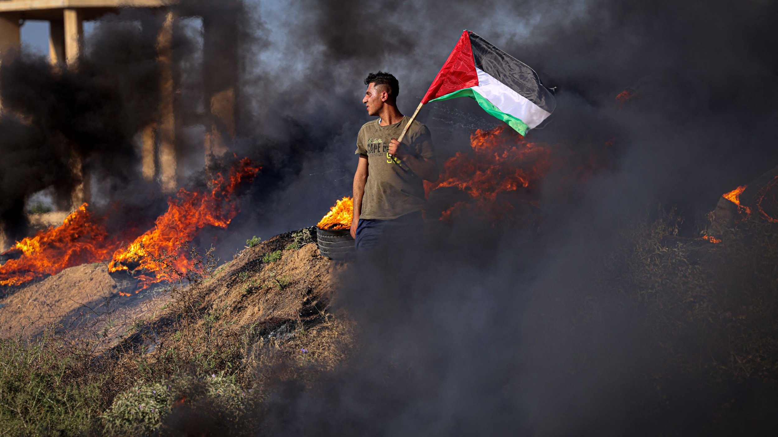تظهر الصورة شابًا يحمل علم فلسطين، يقف وسط دخان كثيف وحرائق مستعرة. يبدو أن الخلفية تعكس حالة من التوتر أو الصراع، حيث النيران تشتعل بالقرب منه. يرتدي الشاب زيًا عاديًا، مما يشير إلى أنه في أوضاع ميدانية. تعكس الصورة مشاعر العزة والانتماء الوطني.