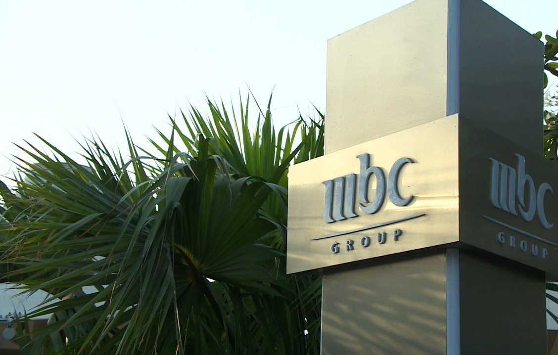 تظهر الصورة لافتة تحمل شعار "مجموعة MBC"، وهو معدني وأنيق، ويقع ضمن بيئة نباتية تتضمن أوراق نخيل وشجيرات خضراء. يبدو أن الصورة التقطت في النهار، حيث يظهر ضوء الشمس بشكل جيد. تعكس الألوان والمواد المستخدمة في التصميم الاحترافية والرقي.