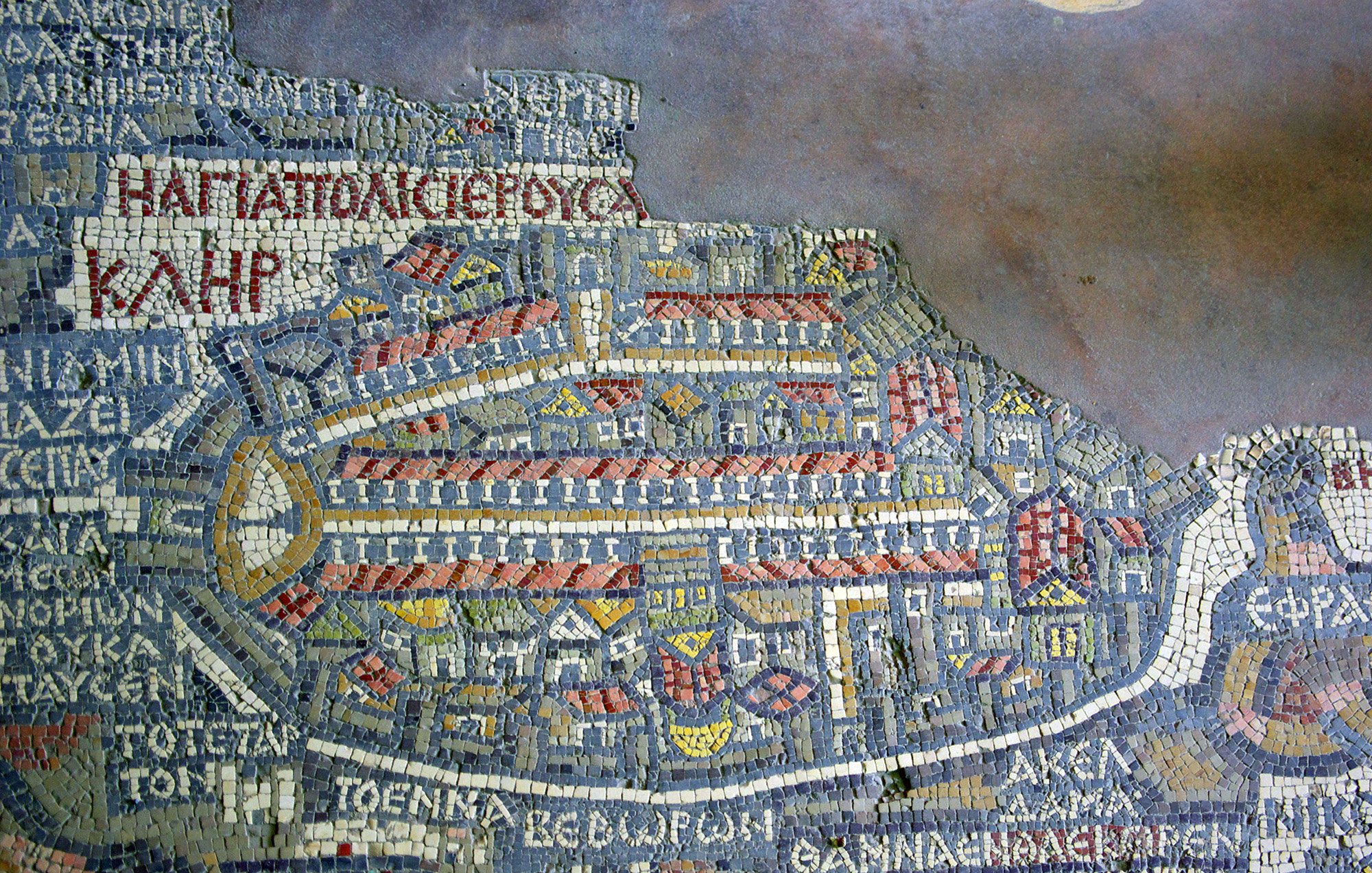 الصورة تظهر فسيفساء تاريخية تمثل خريطة لمكان ما، حيث تحتوي على تفاصيل معمارية وشوارع، بالإضافة إلى نصوص مكتوبة باللغة اليونانية. يظهر فيها تصميم معقد يوضح ملامح المدينة، بما في ذلك الأبنية والمنافذ الرئيسية. الألوان المستخدمة تشمل الأحمر والأزرق والأبيض، مما يضفي جمالية على العمل الفني. هذه الخريطة تعكس التاريخ والثقافة المعمارية للمنطقة التي تمثلها.