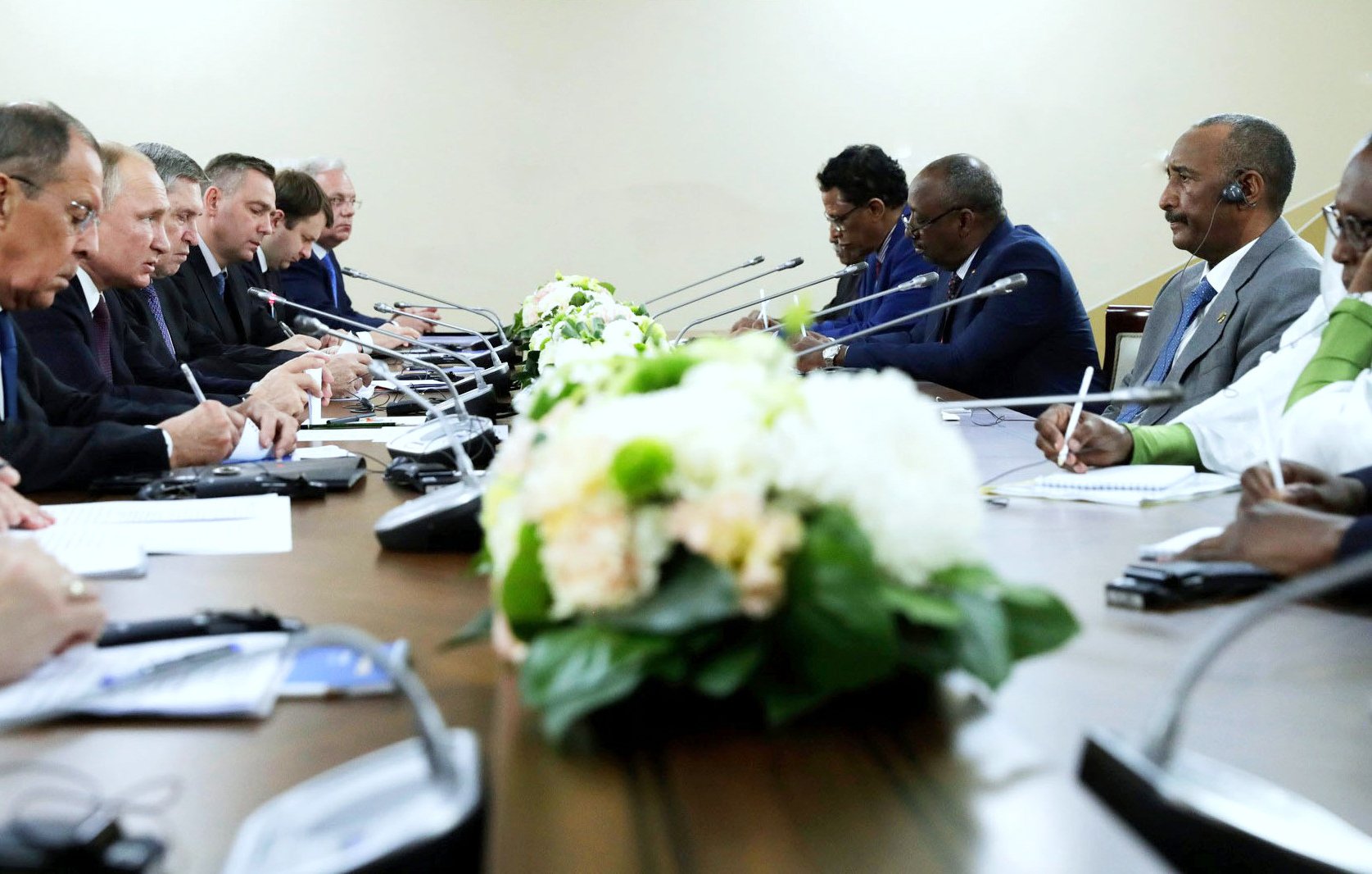 تظهر الصورة اجتماعاً يجري في قاعة مؤتمرات، حيث يجلس عدد من الشخصيات حول طاولة مستديرة. يبدو أن المناقشات جادة، حيث يركز المشاركون على الأوراق أمامهم. يوجد في وسط الطاولة تنسيق للزهور، مما يضيف لمسة جمالية للمكان. المشاركون يرتدون ملابس رسمية، ويبدو أن هناك تبادل للأفكار والمعلومات.