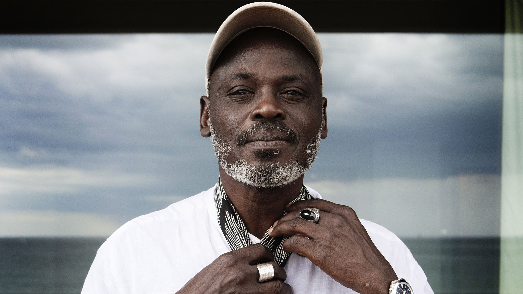 تظهر الصورة رجلًا يرتدي قميصًا أبيض وسلسلة ويضع قبعة على رأسه. يبدو أنه يثبت وشاحًا حول رقبته، ويقف أمام منظر طبيعي يضم البحر في الخلفية. السماء تظهر غائمة وكأنها تحمل بعض الغيوم. ملامح وجهه تعكس الثقة والهدوء.