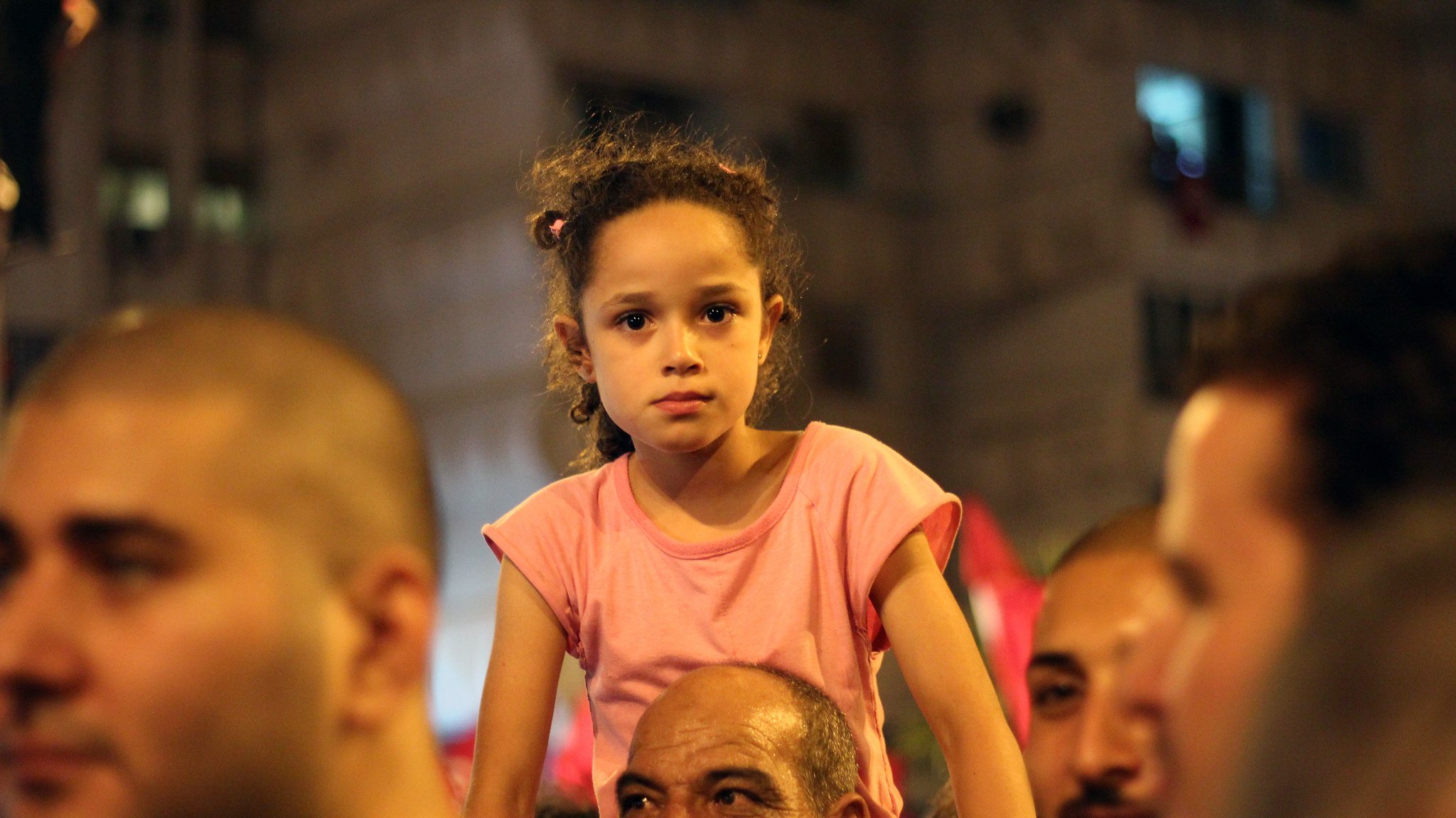 في الصورة نرى مجموعة من الأشخاص في تجمع جماهيري، تركز الكاميرا على فتاة صغيرة تجلس على أكتاف رجل. تبدو الفتاة بملابس بسيطة، ولديها شعر مجعد، وتعبر ملامح وجهها عن اهتمام أو شغف بما يحدث حولها. توجد تعابير متنوعة على وجوه الحضور، مما يعكس الأجواء الاحتفالية أو المتحمسة في هذا الحدث. الب背景 يبدو حضريًا مع مبانٍ مضاءة.