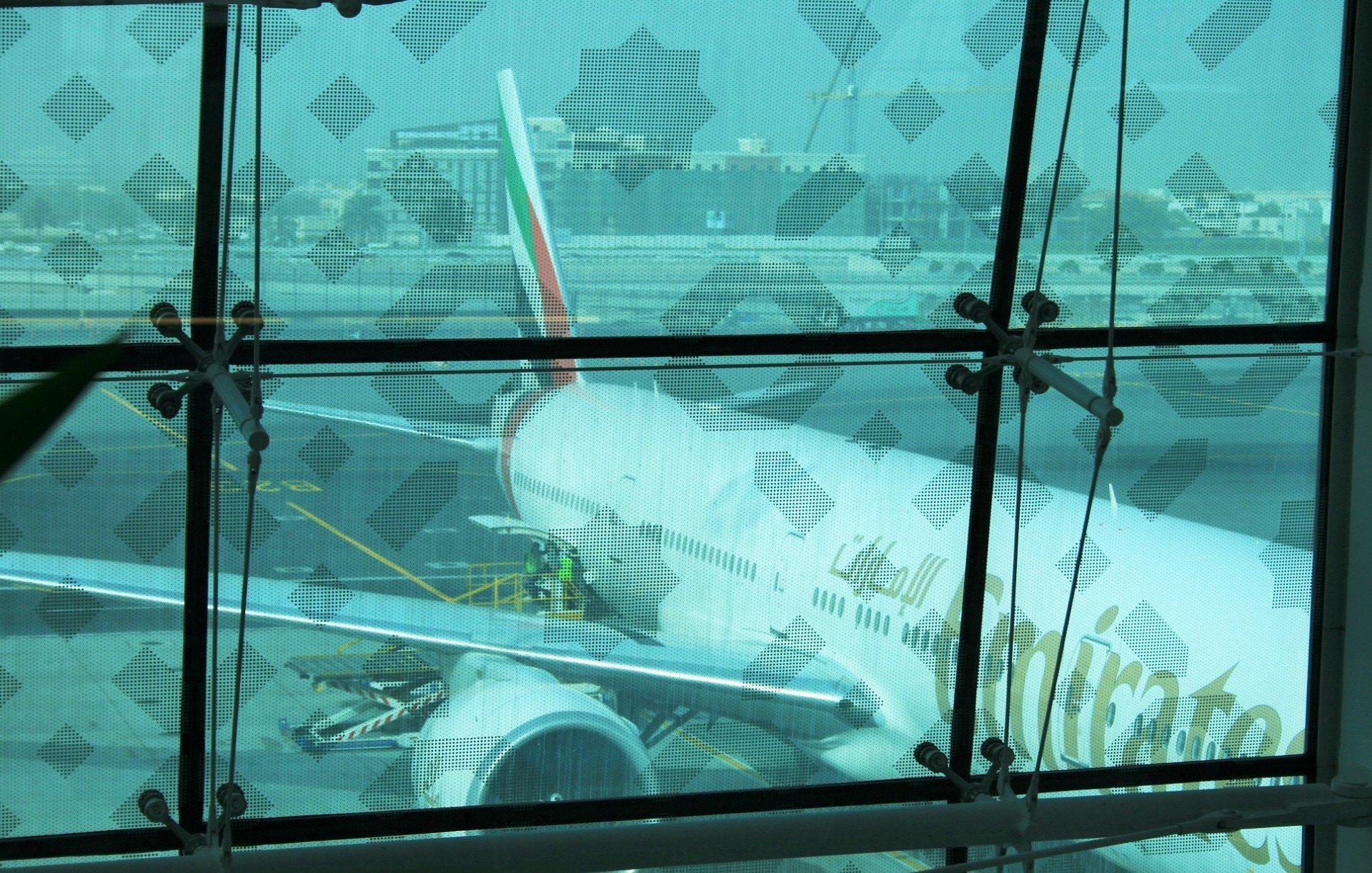 الصورة تظهر مطارًا، حيث يمكن رؤية طائرة تحمل شعار خطوط الإمارات. الطائرة متوقفة على المدرج، ويظهر جزء من هيكلها وأحد محركاتها. خلف الزجاج، يمكن رؤية بعض الأنشطة في المطار، مثل التحميل أو الصيانة، مع وجود بعض العمال في الصورة. الزجاج يحتوي على نمط معين يكرّس الخصوصية. الجو يبدو ضبابيًا بعض الشيء، مما يعكس أوقاتًا مختلفة في المطار.