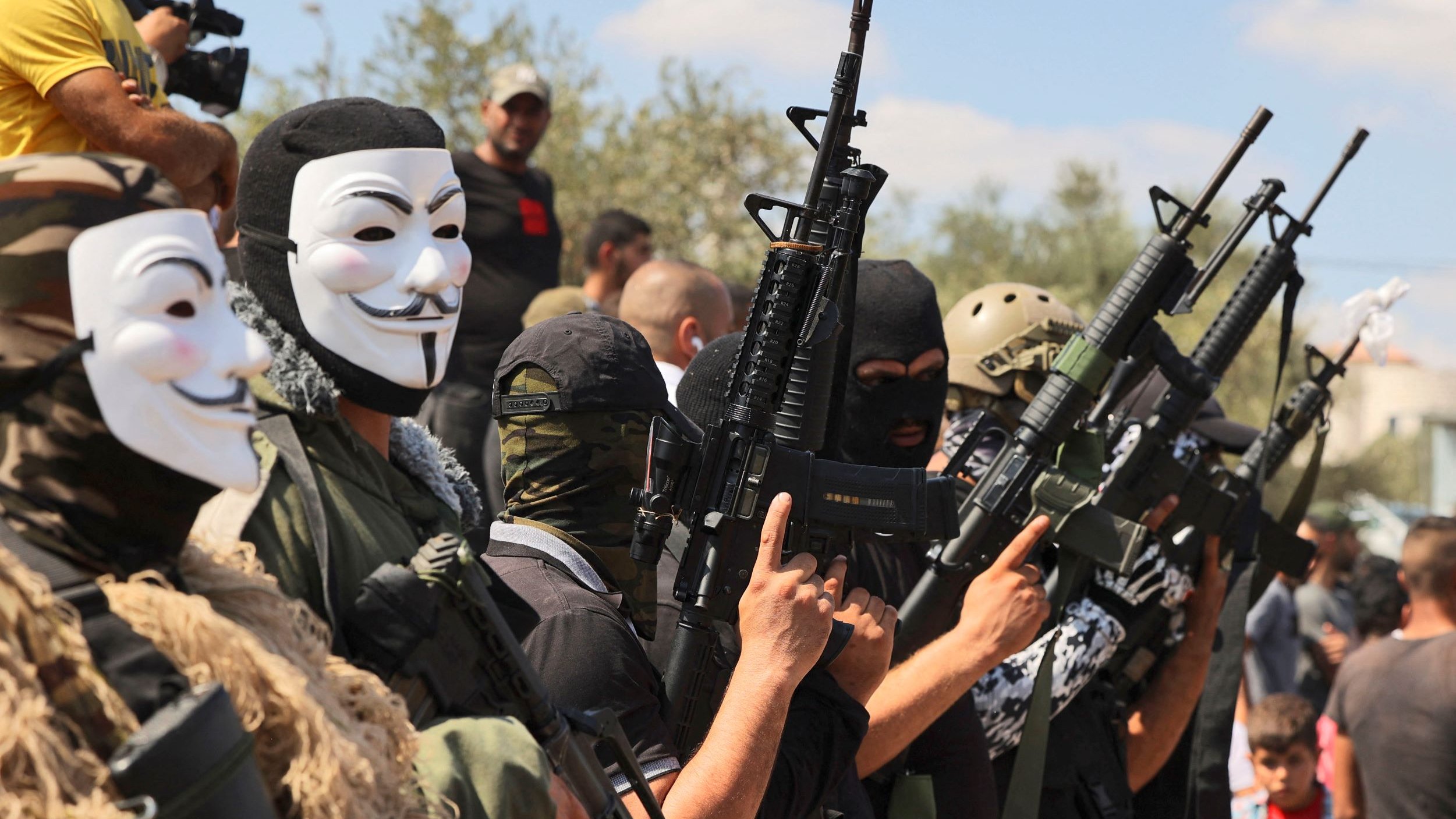 تظهر في الصورة مجموعة من الرجال يرتدون أقنعة وأزياء عسكرية، ويحمون أسلحة متنوعة. خلفهم يوجد عدد من الأشخاص في الخلفية. تعكس الصورة أجواء عسكرية أو استعراض لمجموعة مسلحة، حيث يتم التركيز على أسلحتهم وتنسيقهم. السماء تبدو زرقاء مع بعض السحاب، مما يعطي انطباعًا عن الأجواء المحيطة.