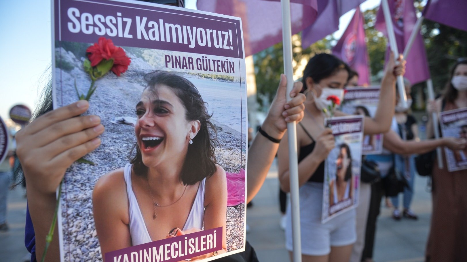تظهر الصورة مجموعة من النساء أثناء تظاهرهن. تحمل إحداهن لافتة مكتوب عليها "Sessiz Kalmıyoruz!" (لا نصمت!) وصورة لامرأة تُدعى "Pınar Gültekin". كما تحمل إحدى المتظاهرات زهرة حمراء، مما يشير إلى أن المظاهرة تتعلق بقضية اجتماعية أو حقوق النساء. يُظهر المشهد روح التضامن والمطالبة بالعدالة.