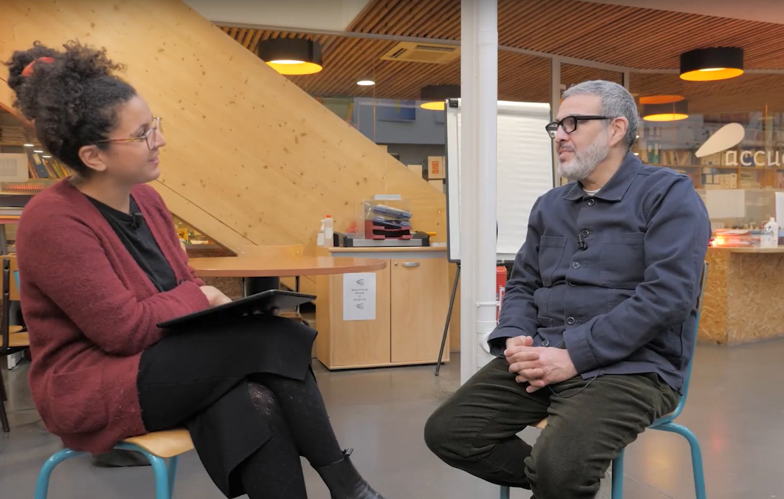 تظهر الصورة مشهداً يضم شخصين يتحدثان معاً في مكان داخلي. الشخص الجالس هو رجل يرتدي قمصاناً داكنة، بينما المرأة تجلس أمامه تحمل جهازاً لوحياً وتبدو وكأنها تستمع إليه. الخلفية تحتوي على رفوف تحتوي على كتب وبعض الأثاث الخشبي. الإضاءة في المكان جيدة، مما يضيف جواً مريحاً للمحادثة.