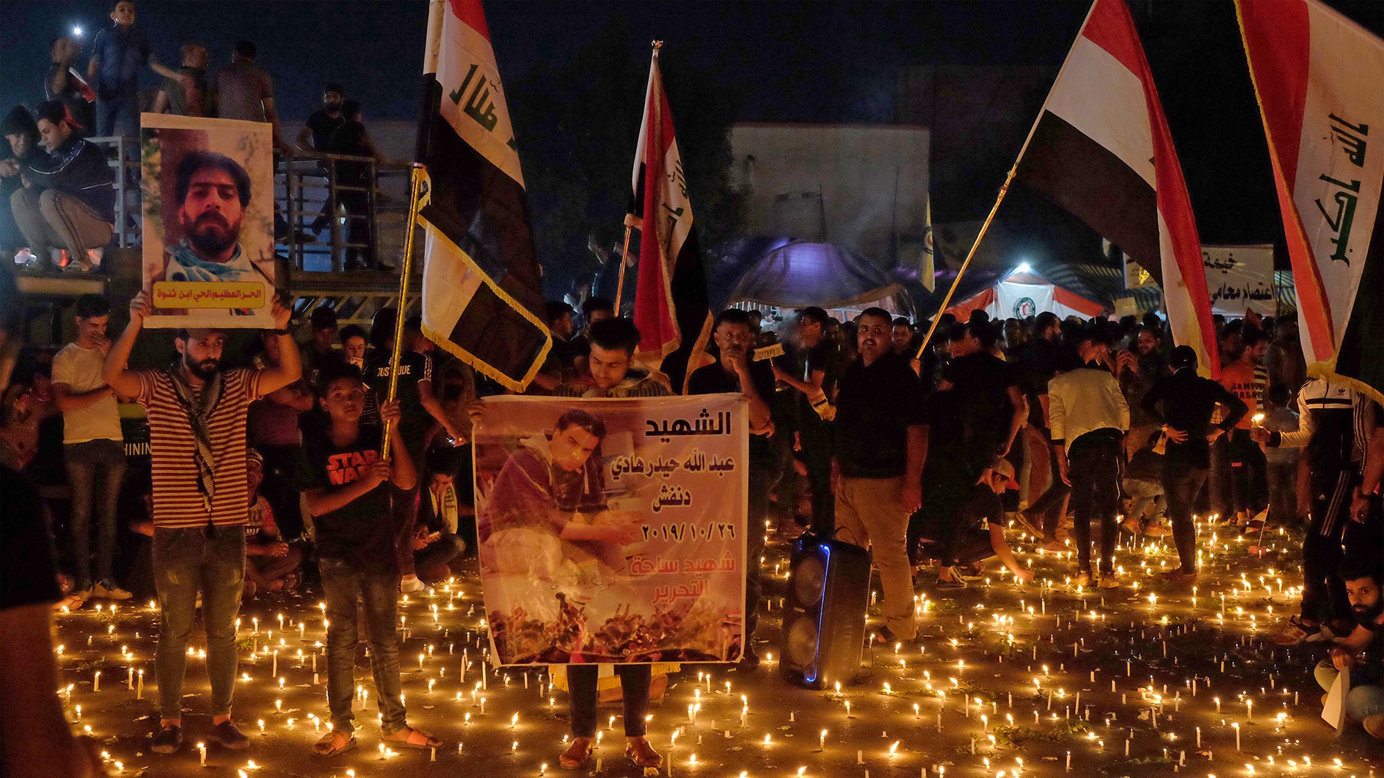 توضح الصورة تجمعًا حاشدًا للناس في مكان مفتوح خلال الليل، حيث يضيء المكان بكثرة الشموع الموزعة على الأرض. يحمل المشاركون لافتات وصورًا، وبعضهم يرفع أعلامًا تمثل بلدانهم. الجو يبدو مهيبًا، حيث يعبر الناس عن حزنهم أو إحياء ذكرى معينة، مما يعكس شعورًا بالوحدة والتضامن في هذه اللحظة.