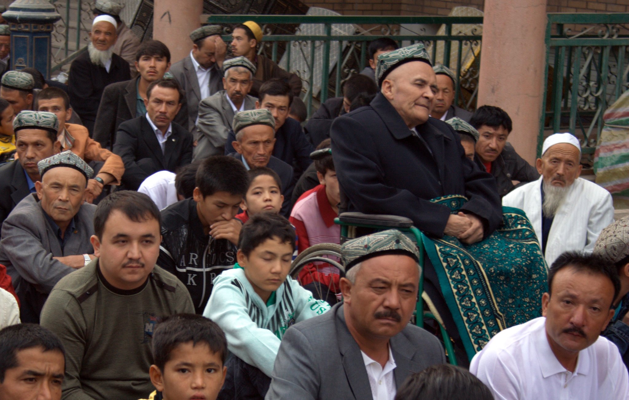 تظهر الصورة مجموعة من الأشخاص الجالسين في مكان مفتوح، يبدو أنهم مجتمعون في مناسبة أو تجمع. يوجد رجل مسن يجلس على كرسي متحرك في الخلف، ويبدو أنه يراقب ما يحدث من حوله. هناك تنوع في الأعمار بين الحضور، حيث توجد وجوه مراهقين وشباب وكبار في السن. يرتدي الكثير من الرجال قمصاناً تقليدية، وبعضهم يرتدي قُبُعات ذات تصاميم مميزة. يبدو أن الجو يسوده الانتباه والتركيز على الحدث الجاري.