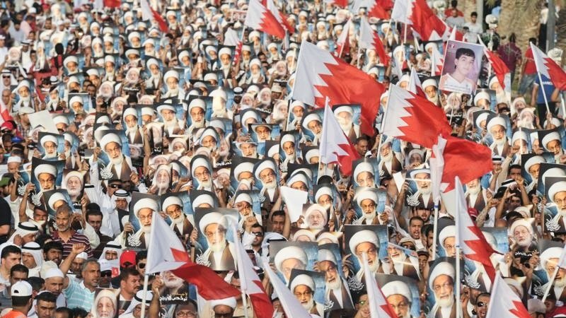 الصورة تُظهر تجمعاً حاشداً لمتظاهرين في البحرين. المشاركون يحملون أعلام البحرين، بالإضافة إلى صور لشخصيات معروفة. الجو يبدو حماسياً، حيث يرتدي العديد من المشاركين الثياب التقليدية. المناظر تعكس روح التضامن والمشاركة الفاعلة في الحدث.