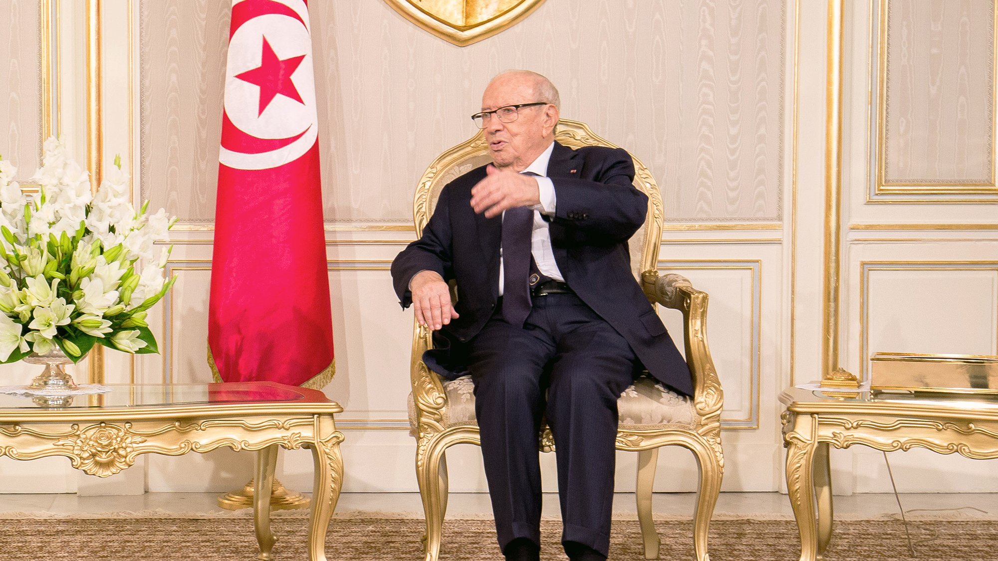 الصورة تظهر رجلًا يجلس على كرسي فاخر في غرفة مزينة بشكل أنيق. خلفه علم تونس، وهناك طاولة صغيرة أمامه تحتوي على زهور بيضاء. يبدو الرجل في وضع يتحدث أو يشرح شيئًا، ويرتدي بدلة رسمية وقميص. الغرفة تبرز بديكوراتها المثالية وألوانها الهادئة.