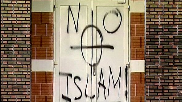 تظهر في الصورة بابًا مغطى بكتابات جدارية، حيث تم رسم عبارة "لا إسلام" مع رمز يظهر قوسًا دوائريًا. الكتابة تبدو باللون الأسود على خلفية بيضاء. الجدران المحيطة بالباب مصنوعة من الطوب بلون بني فاتح. الصورة تعكس مشاعر سلبية تجاه الإسلام، مما يثير جدلاً حول التسامح والقبول.