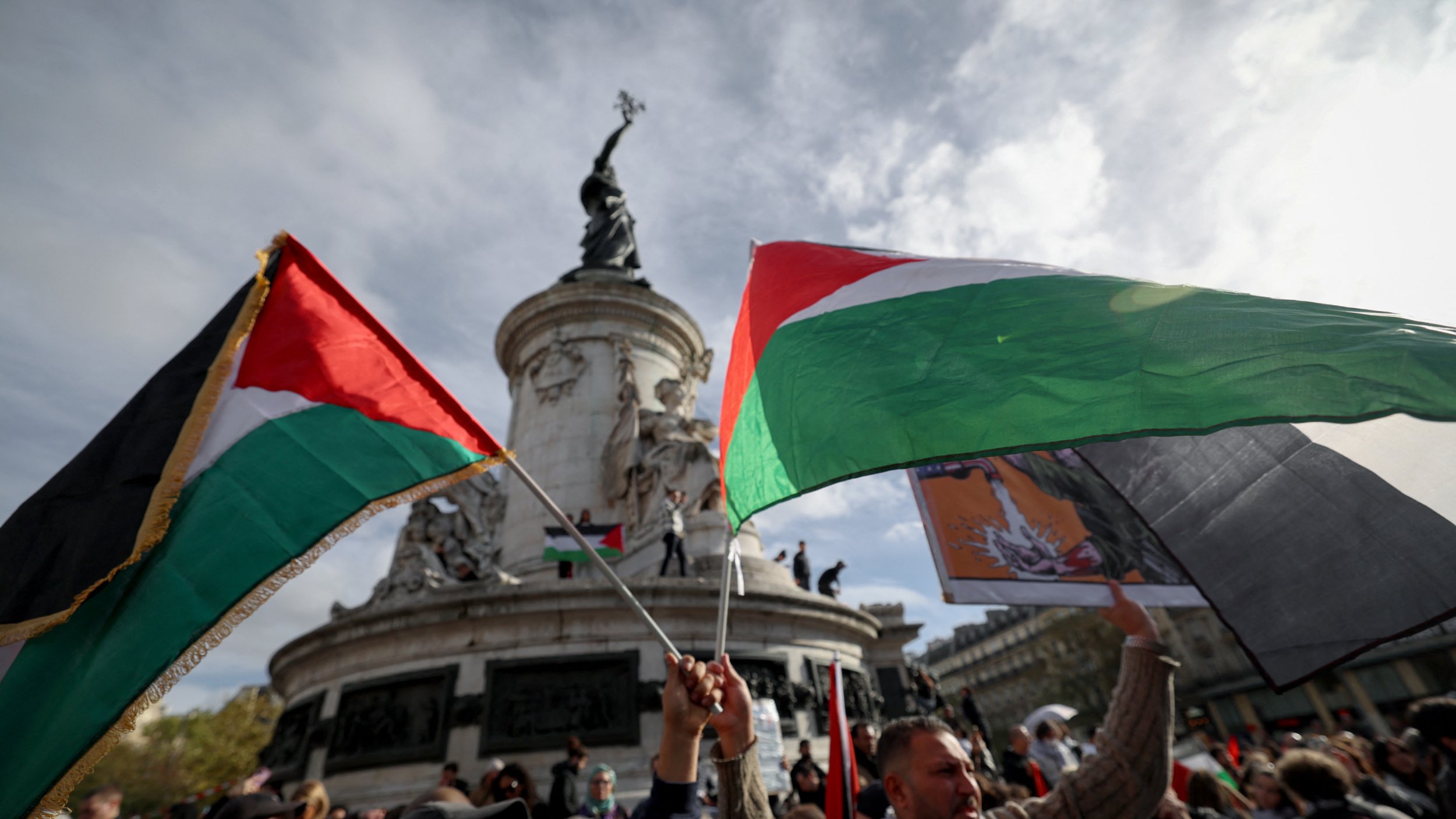 تظهر الصورة مجموعة من الأشخاص وهم يحملون أعلام فلسطينية في تجمع جماهيري. الخلفية تُظهر نصبًا تذكاريًا عظيمًا، مع سماء ملبدة بالغيوم. يبدو أن الناس متحمسون ويشاركون في فعالية ذات طابع وطني أو سياسي. الأعلام تحمل ألوان العلم الفلسطيني، مما يشير إلى دعمهم للقضية الفلسطينية.