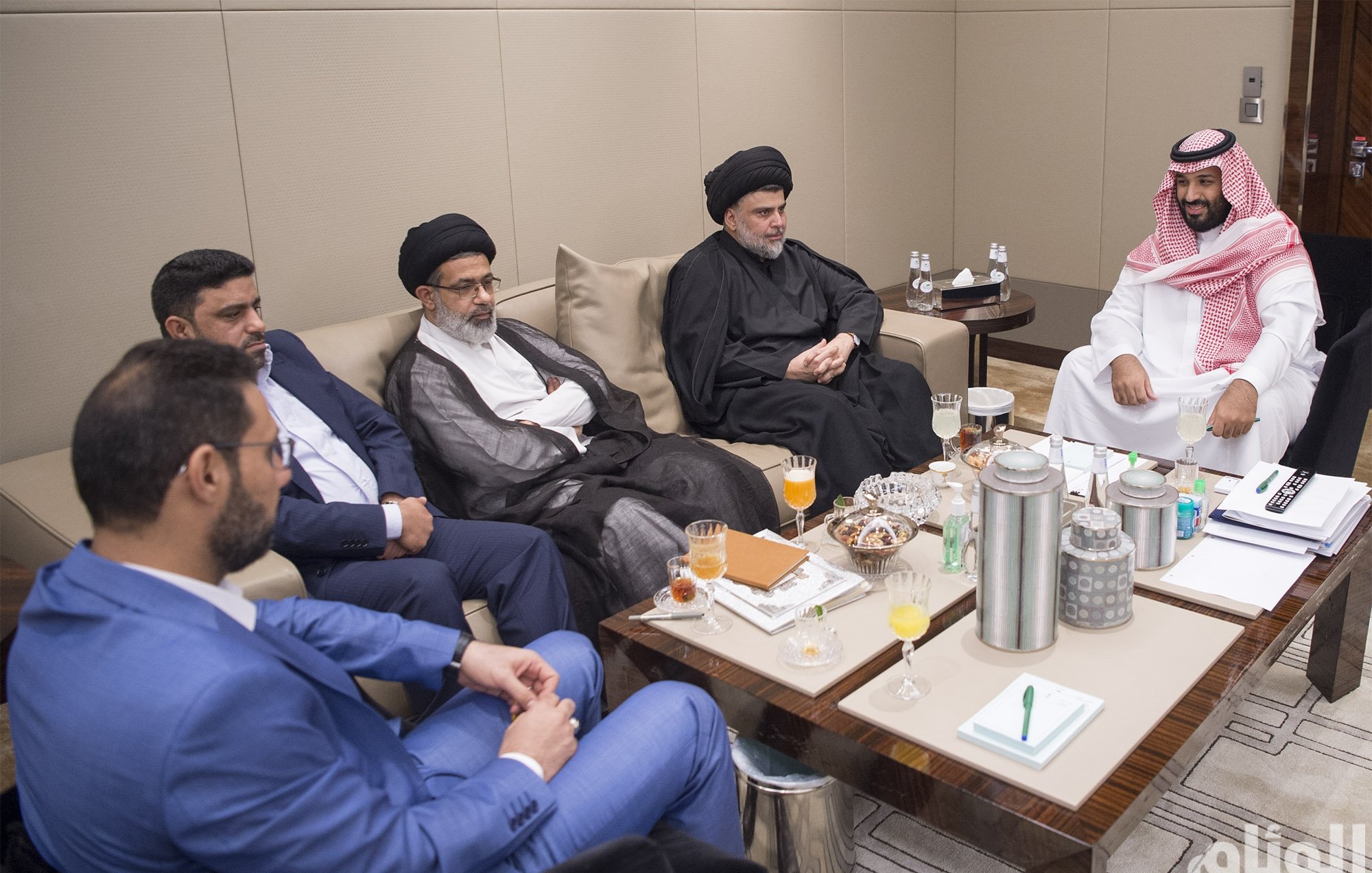 تظهر الصورة مجموعة من الأشخاص يجلسون في قاعة اجتماعات. يتواجد بعضهم بملابس تقليدية، بينما يرتدي آخرون ملابس رسمية. على الطاولة أمامهم توجد بعض المشروبات والوجبات الخفيفة. يبدو أن الاجتماع يتضمن مناقشات بين الأفراد، حيث يمتلك كل منهم تعبيرات جدية. المكان يبدو أنيقًا وعصريًا، مما يوحي بأهمية اللقاء.