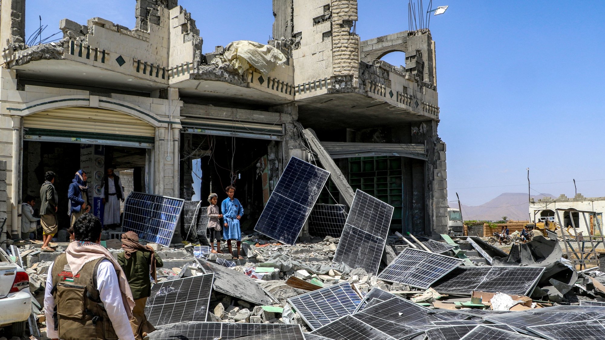 صورة لمكان مدمر، مباني مهدمة وألواح شمسية متكسرة، وأشخاص يتجمعون في الموقع.