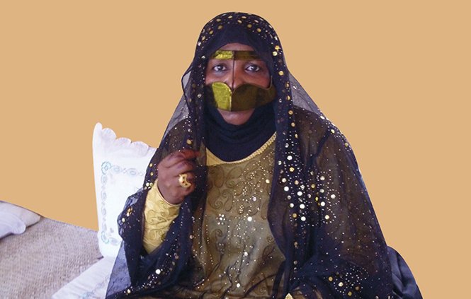 الصورة تظهر امرأة ترتدي زيًا تقليديًا مميزًا، والذي يتضمن عباءة سوداء تغطي جسدها وزينتها بألوان ذهبية أو لامعة. وجهها مغطى جزئيًا بحجاب، مما يبرز فقط عينيها. المرأة تجلس في بيئة منزلية بسيطة، حيث توجد بعض الأواني والزينة حولها. تعكس الصورة تراثًا ثقافيًا غنيًا وتفاصيل تقليدية لعادات معينة.