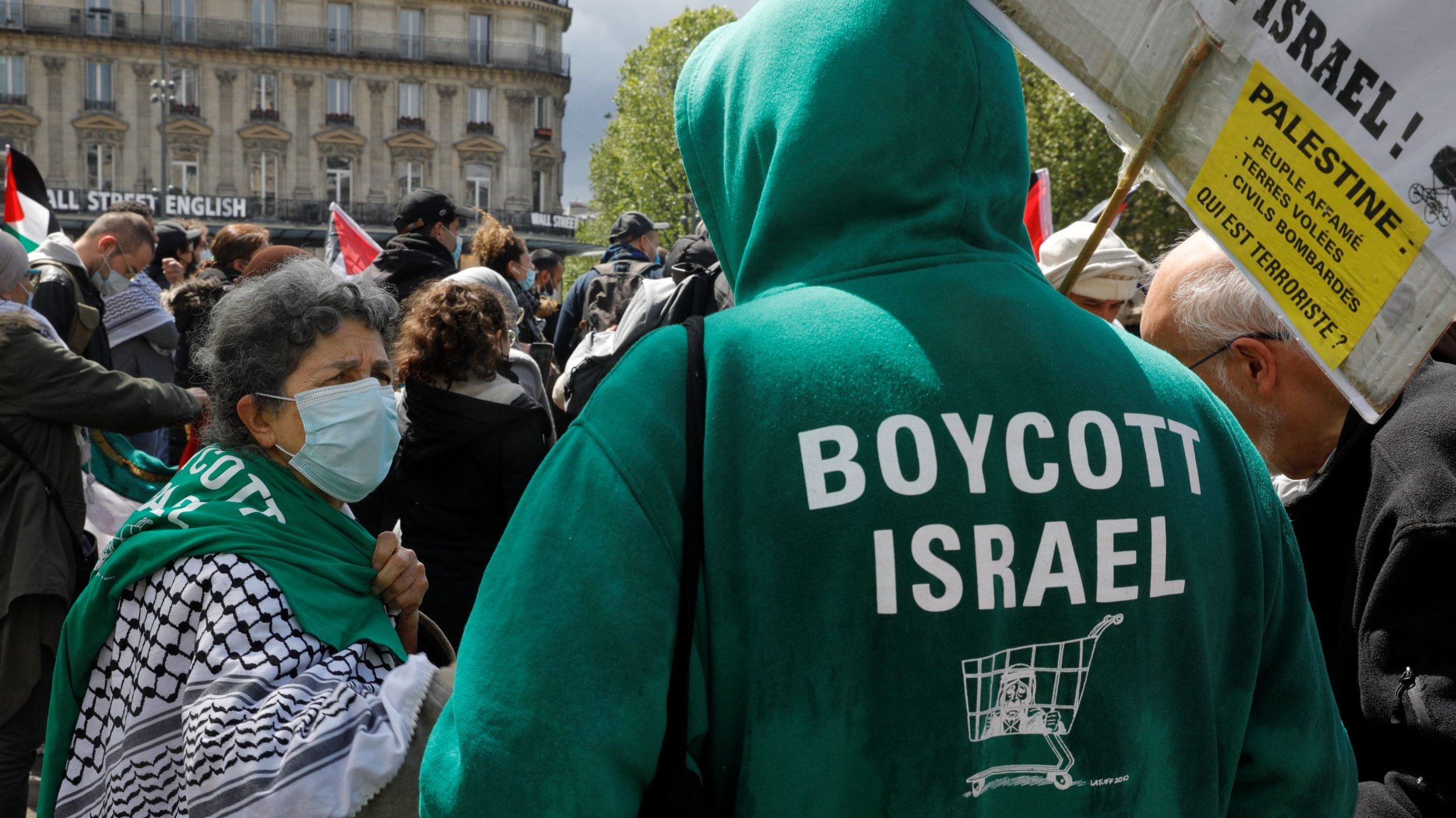 تظهر في الصورة مجموعة من الأشخاص في تظاهرة تتعلق بقضية فلسطين. يرتدي أحد المتظاهرين سترة خضراء مكتوب عليها "BOYCOTT ISRAEL"، مما يعبر عن دعوة لمقاطعة إسرائيل. في الصورة أيضًا، هناك امرأة ترتدي كمامة وتبدو أنها تتحدث مع المتظاهرين الآخرين. الخلفية تشمل مباني ورايات تحمل شعارات مختلفة. الجو العام يوحي بالتضامن والمشاركة في حركة مناهضة للاحتلال.