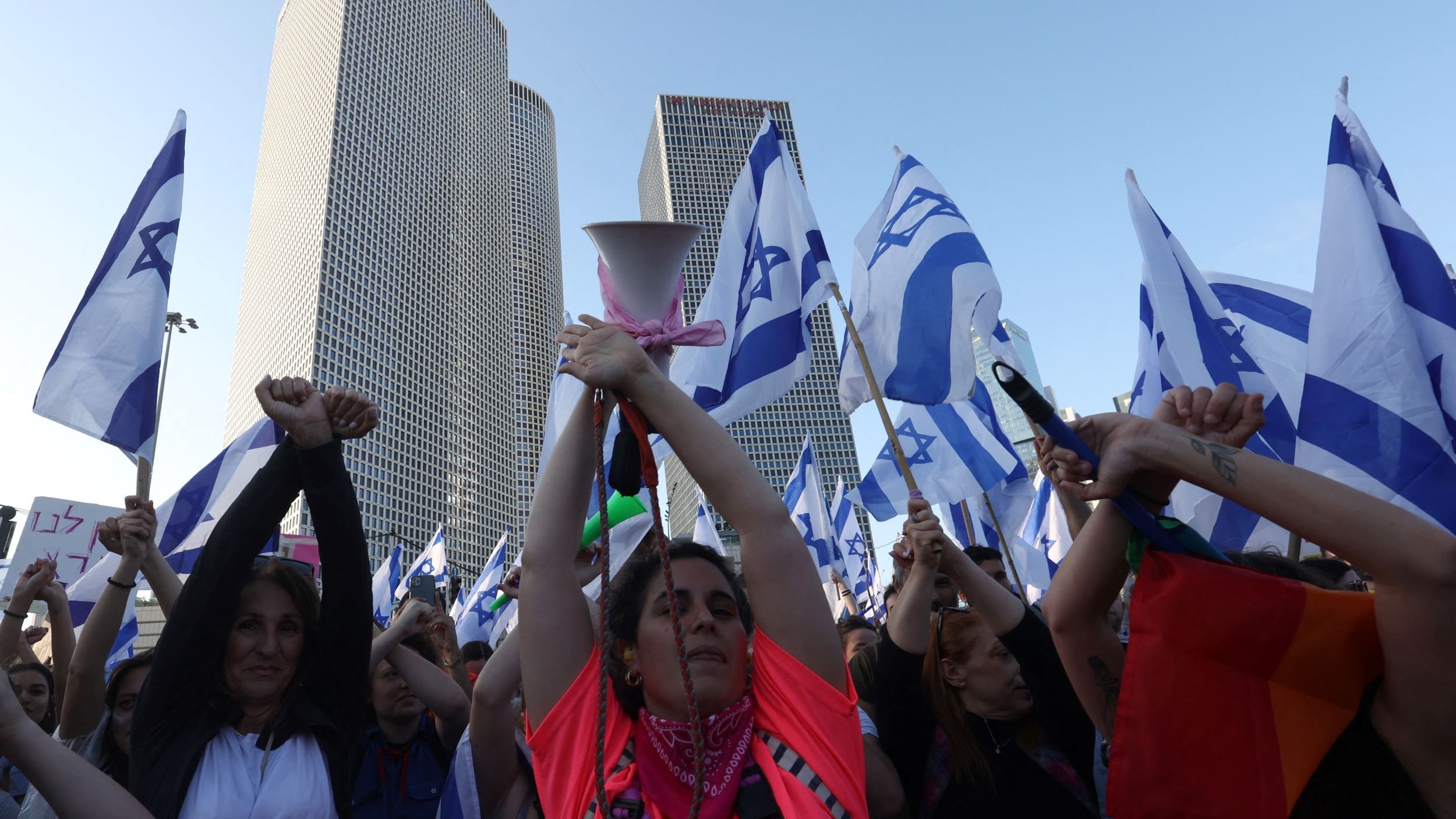 تظهر الصورة مجموعة من الأشخاص في مظاهرة، يحملون أعلامًا تحمل نجمة داود، وهو رمز معروف في إسرائيل. تظهر امرأة في المركز، مرفوعة اليدين وتحمل جهاز مكبر صوت، بينما يرفع الآخرون الأعلام. تتواجد خلفهم بنايات حديثة، مما يشير إلى أن المظاهرة قد تكون في منطقة حضرية. يعبّر المتظاهرون عن آرائهم، مما يدل على أهمية الفعالية التي يشاركون فيها.