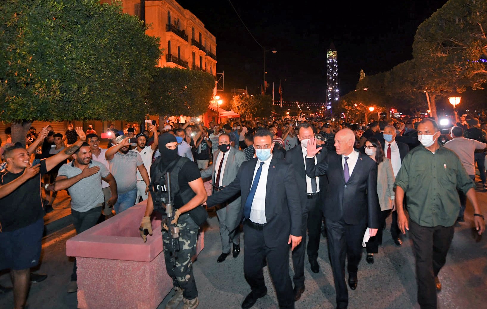 تظهر الصورة مجموعة من الأشخاص في شوارع مدينة مضاءة بأضواء ليلية. يتواجد في المقدمة رجل يرتدي بدلة مع أشخاص آخرين، من بينهم رجل ملثم يحمل سلاحًا. يبدو أن هناك تجمعًا من الحشود في الخلفية، حيث يتفاعل بعضهم مع الموقف. الأجواء توحي بوجود حدث أو مظاهرة، ومشاعر الحماس والانفعال واضحة في تعابير الحضور.