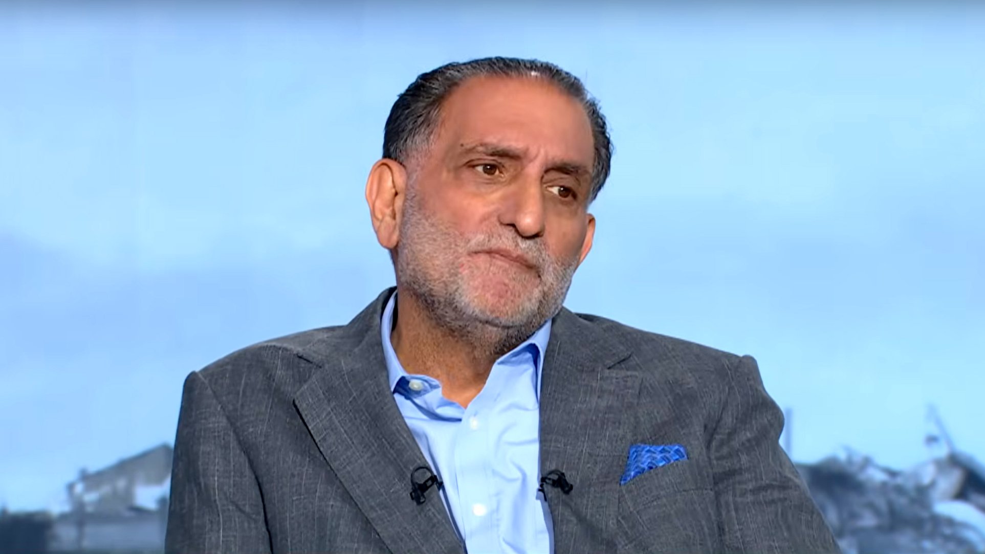 رجل يجلس مرتديًا بدلة رمادية، يبدو عليه التفكير والاهتمام.