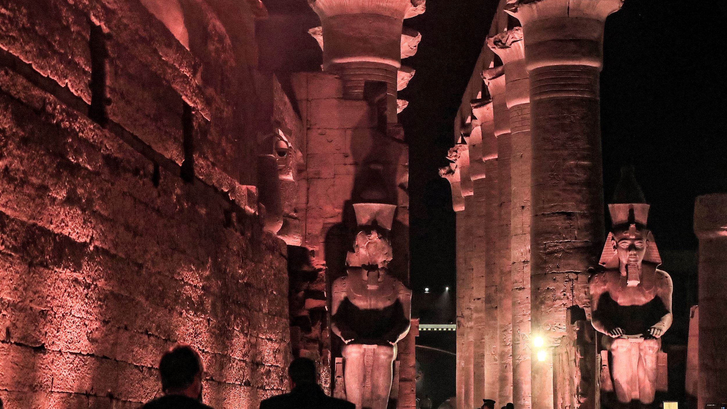 الصورة تظهر معالم أثرية قديمة، حيث يمكن رؤية أعمدة ضخمة ومعمار تاريخي فاخر. الإضاءة الليلية تضفي جوًا سحريًا على المكان، مع تماثيل كبيرة تشكل جزءًا من التصميم المعماري. يبدو أن هناك زوارًا يستكشفون هذا الموقع التاريخي، مما يدل على شهرة وأهمية هذا المكان السياحي.