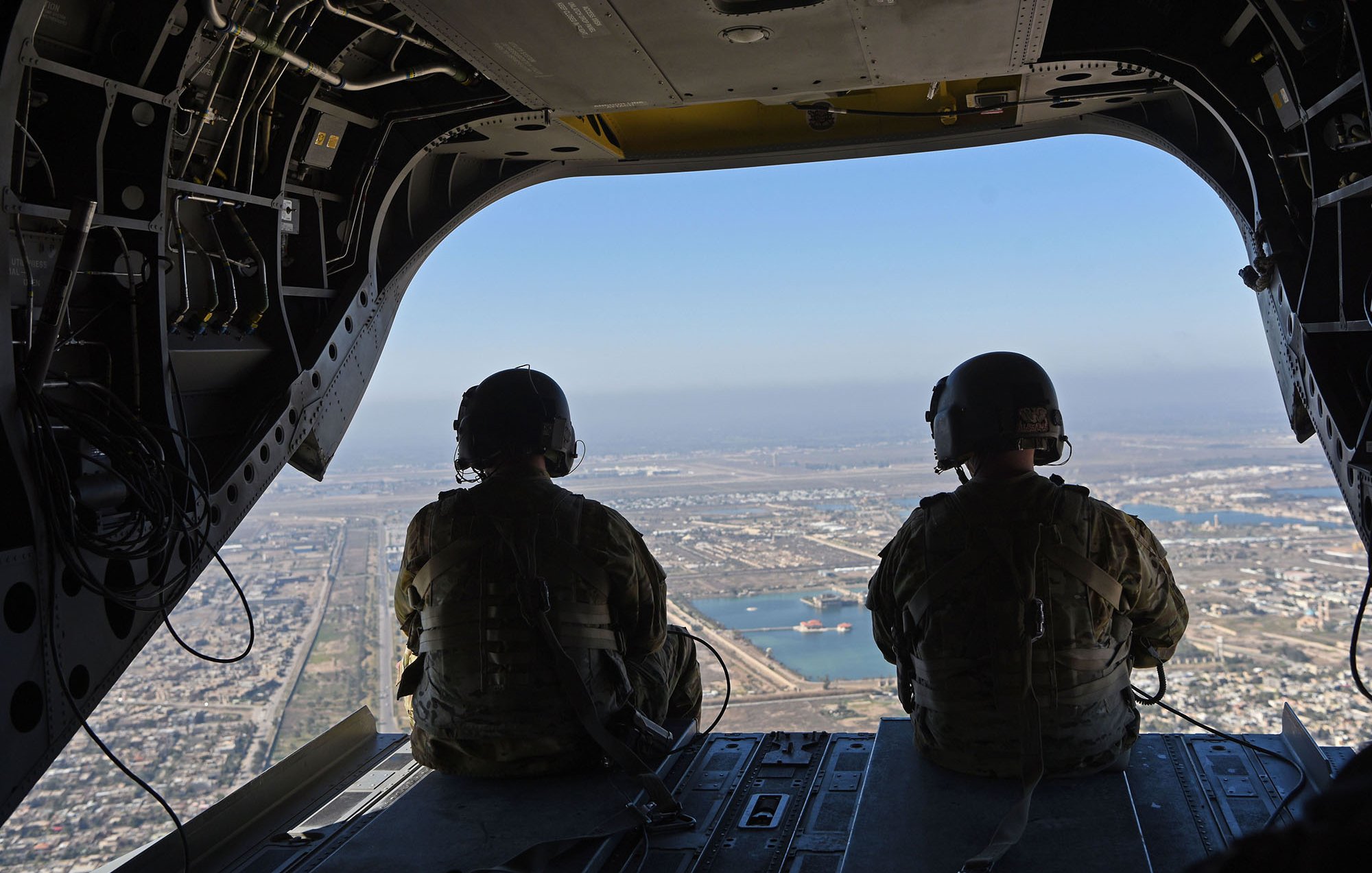 تظهر الصورة مشهداً من داخل طائرة عسكرية، حيث يجلس جنديان يرتديان خوذات وسترات عسكرية. يتم تصويرهما من الخلف وهما يتطلعبان من فتحة الطائرة. يظهر في الأفق منظر للمدينة مع ممرات للمطار وبعض المباني والماء في الخلفية. السماء واضحة وتبدو هادئة.